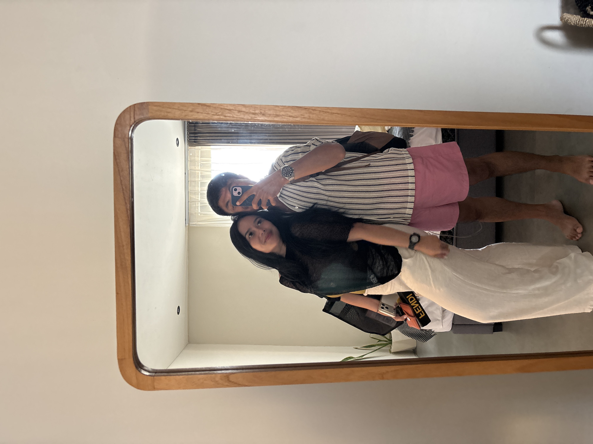 A full-length mirror reflects a couple taking a mirror selfie. The woman, on the left, leans her head on the man's shoulder and smiles. She is wearing a sheer black top and long, loose white pants. She holds a small orange bag with "FENDI" written in yellow text. The man, on the right, wears a striped short-sleeved shirt and pink shorts. He is holding a smartphone to take the picture. The background of the reflection shows a room with a bed, a plant in a pot, and curtains. The location is stated as Mengwi, Indonesia.