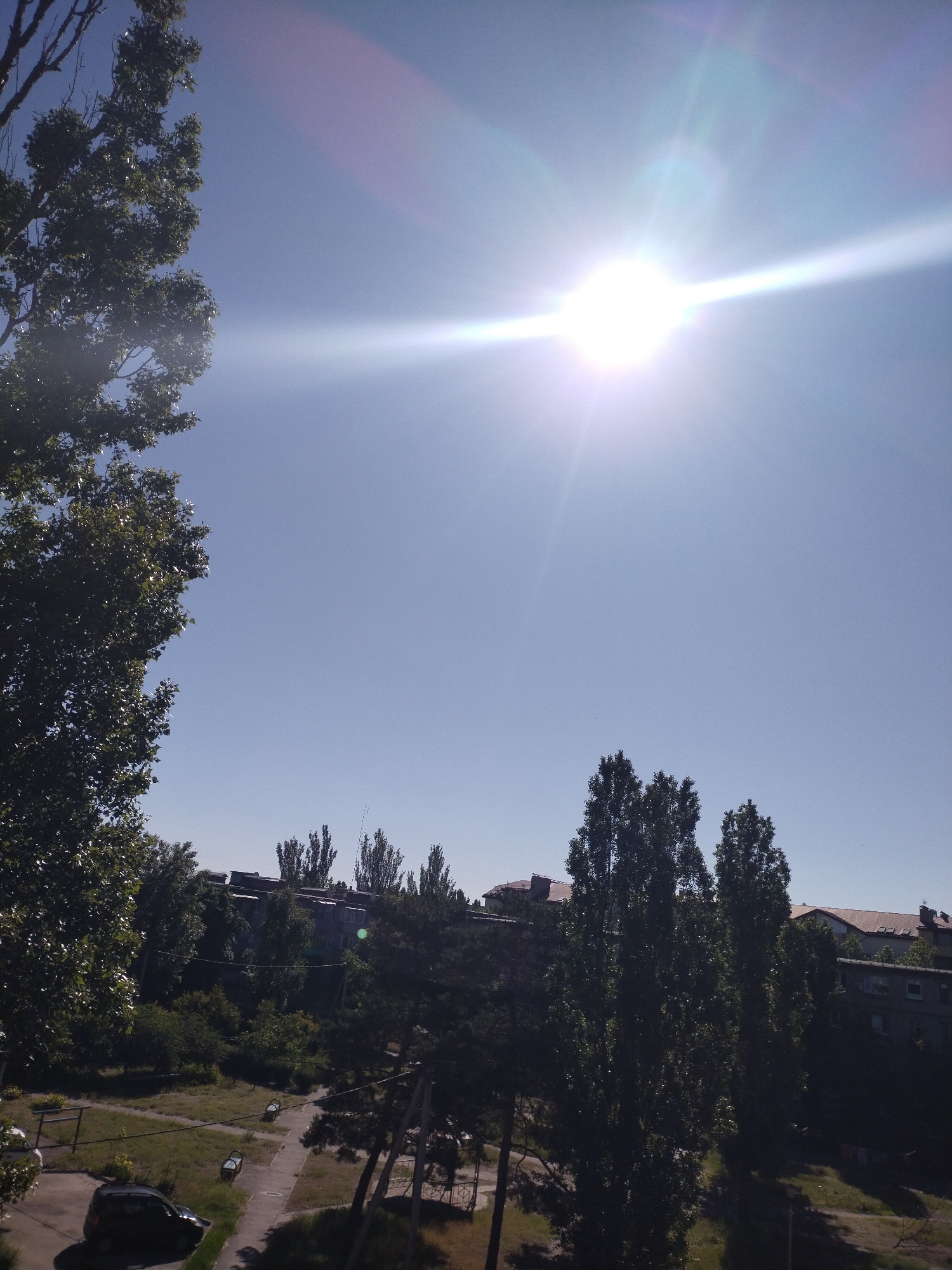 The image shows a view in Horishni Plavni, Ukraine, looking out over a mix of greenery and buildings under a bright, clear sky. The sun is high in the sky, causing a bright glare and lens flare. Trees frame the left and bottom right of the image. A car is parked on a street at the bottom left. In the distance are buildings with varying rooflines and heights, interspersed with tall, slender trees. The scene appears to be taken from a higher vantage point, such as a balcony or upper floor.