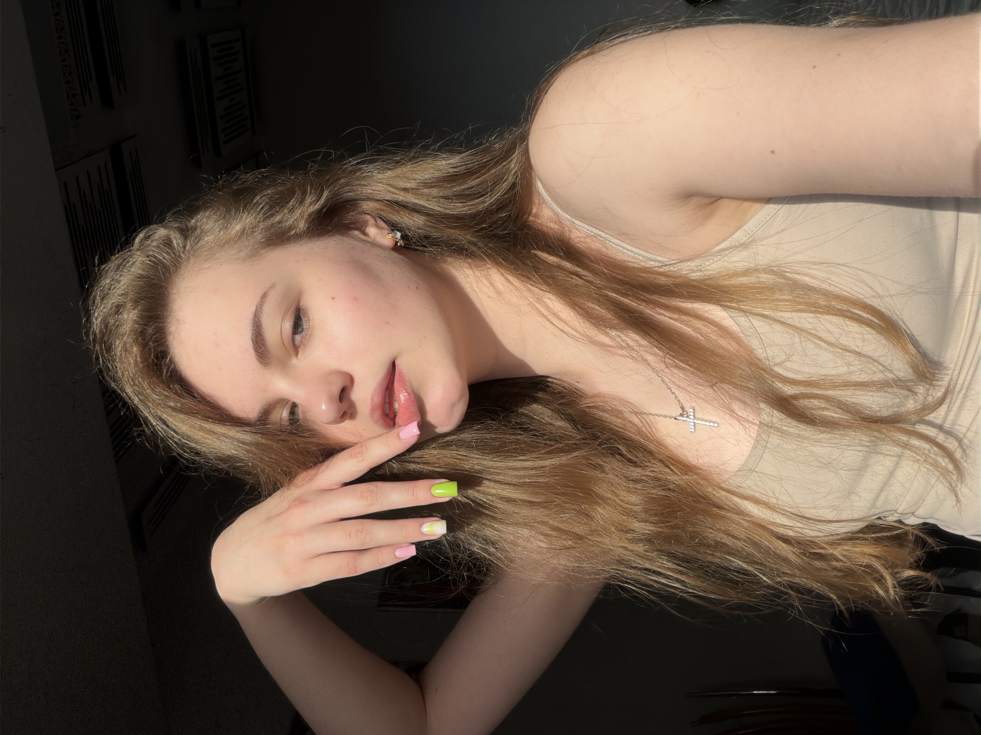 A young woman with long, blonde hair is shown from the chest up, positioned in a way that her right arm is bent and her hand is near her face, obscuring part of her cheek. Her eyes are looking towards the camera. She is wearing a light-colored tank top and a silver cross necklace. Her fingernails are painted with a mix of pink and neon green colors. The lighting suggests a sunlit room, with shadows cast on the wall behind her. The context of Dnipro, Ukraine, is not visually discernible from the image.