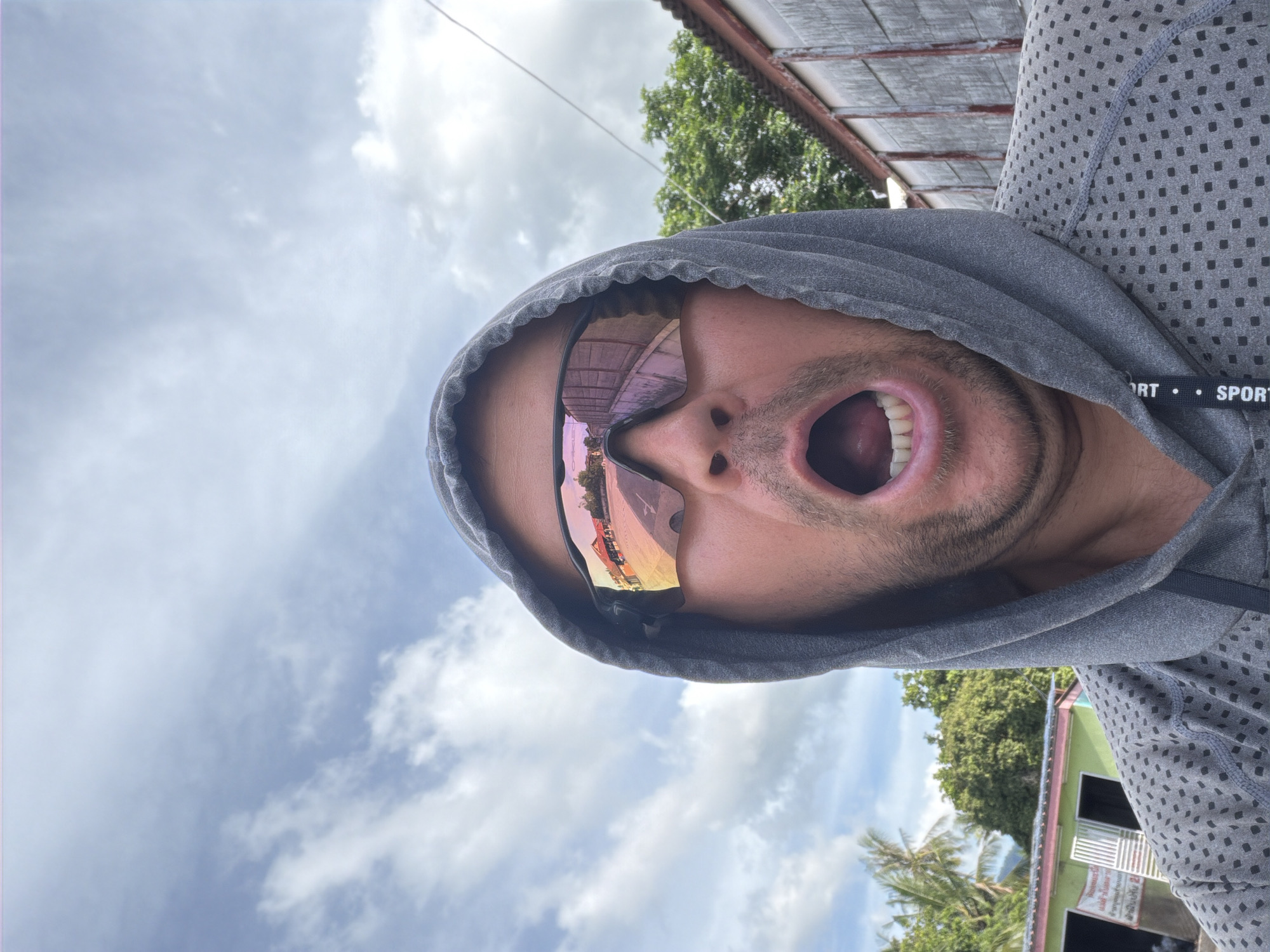 A man wearing a gray hooded sweatshirt and sunglasses is captured in a selfie outdoors. His mouth is open in a surprised expression. The sunglasses reflect a scene with buildings and trees, suggesting an outdoor environment. In the background, there are palm trees and buildings, consistent with a tropical location like Baan Mae Nam, Thailand. The sky is partly cloudy.
