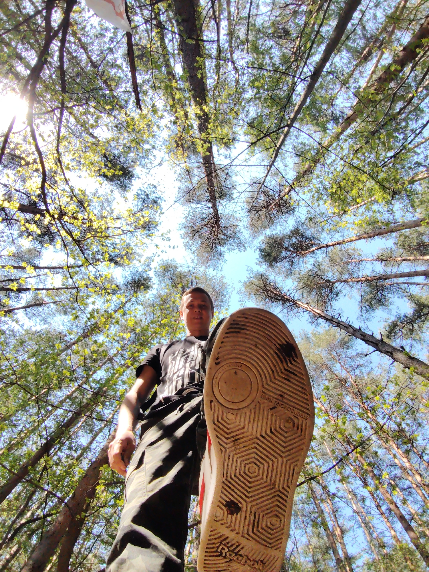 A low-angle shot taken in a forest, possibly in the city of Chaikovsky, Russia, features a person standing and looking down at the camera with one shoe raised, sole facing the viewer. The forest has tall, thin trees reaching upwards, with green leaves and branches visible against a bright, partly cloudy sky. The person is wearing a black t-shirt with a design and dark pants with lighter patches. The sole of the raised shoe is off-white with a geometric, hexagonal pattern and some dirt. The overall composition emphasizes the height of the trees and the perspective from below.
