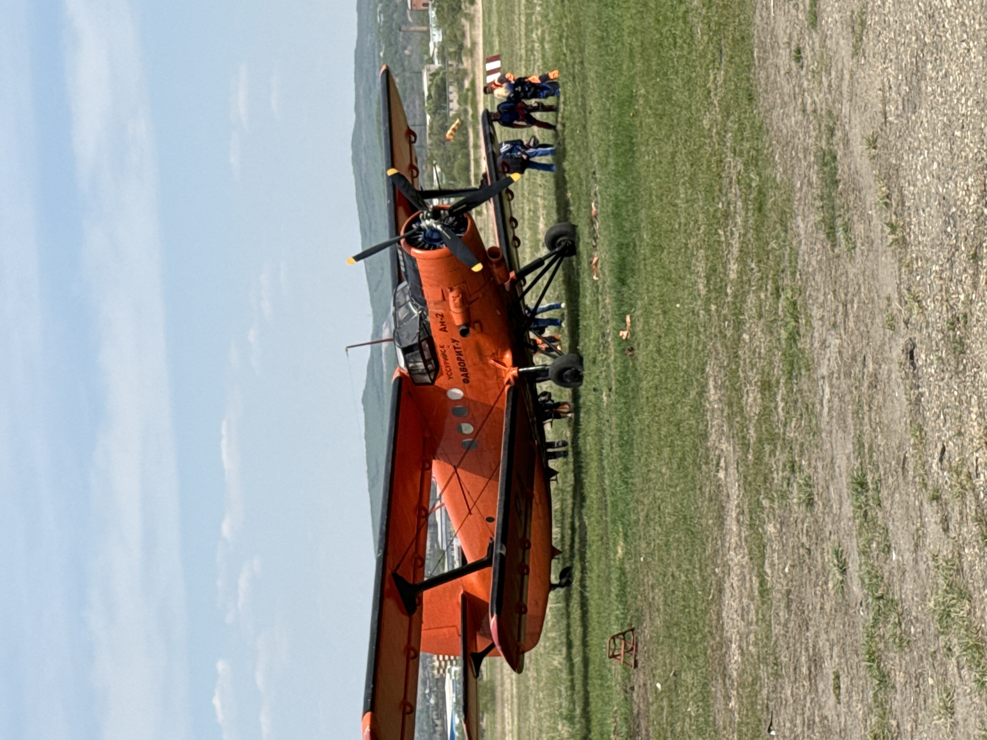 An orange An-2 aircraft is parked on a grassy field in Ussuriysk, Russia, with a group of people gathered near its tail. The biplane has a black propeller and black accents on the wings and fuselage. The text "УССУРИЙСК АН-2 ФАВОРИТ-У" is visible on the aircraft's body. The terrain surrounding the plane appears to be a mix of grassy and gravelly areas, with a building or structure in the distance. The sky is partly cloudy.