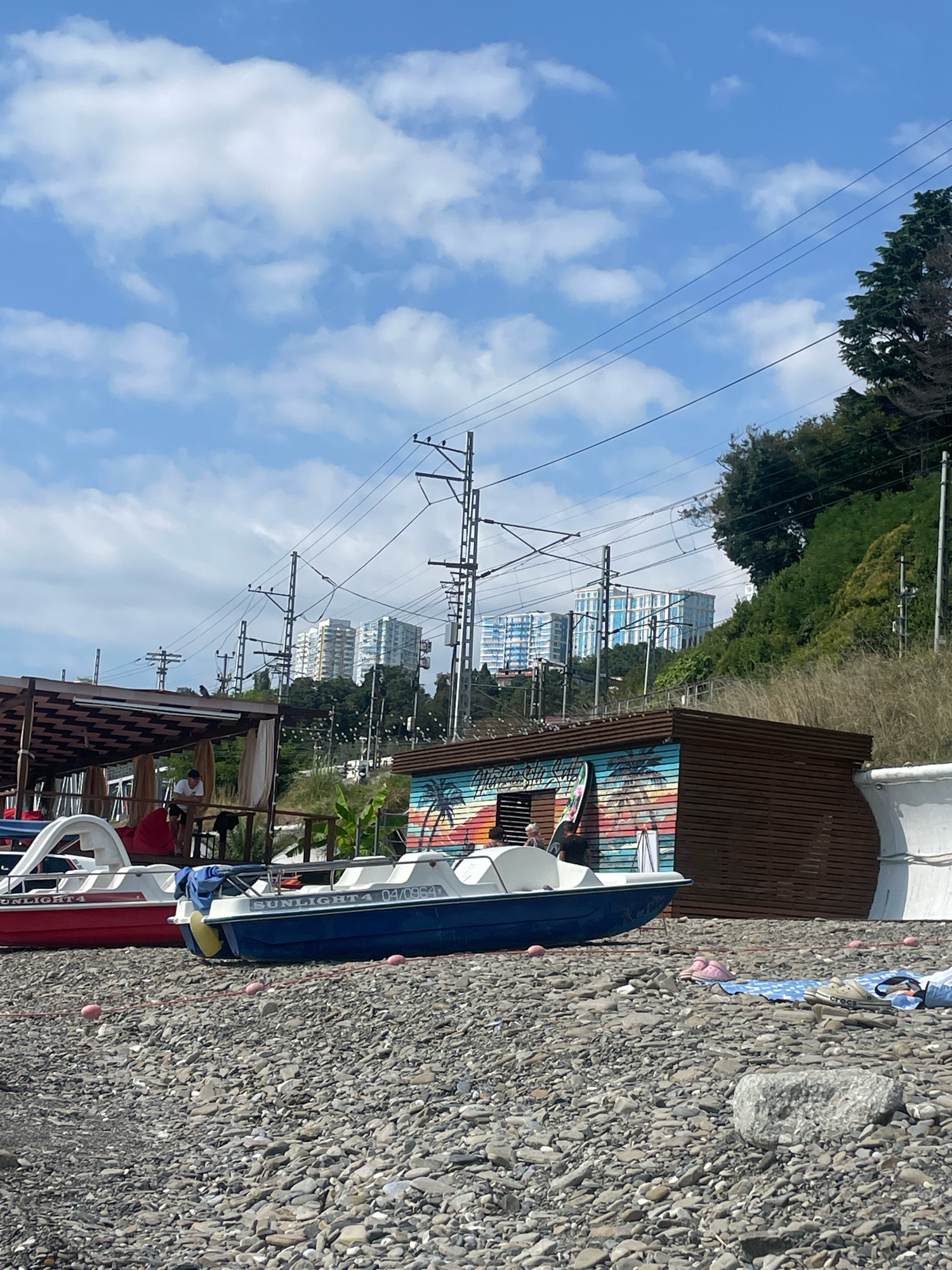 This image captures a seaside scene on a pebble beach in Sochi, Russia, under a partly cloudy sky. In the foreground, two pedal boats are visible on the shore. The boat on the left is red and white, while the one on the right is blue and white with the marking "SUNLIGHT4 04/0964". A person is seated inside the blue boat, and another person is leaning on a railing of a structure behind the boats.
Behind the beach, a rustic wooden structure with a colorful mural depicting palm trees and a sunset is situated. To the left of this structure, a shaded area with a patterned roof and beige curtains provides shelter. Beyond these structures, a steep, grassy hillside is covered with lush green trees. In the distance, several tall, modern apartment buildings with white and blue facades are visible, nestled against the hillside. Overhead, a network of electrical wires and utility poles crisscrosses the sky, indicating the presence of a railway line or power infrastructure. The ground is covered with a dense layer of grey and brown pebbles. A blue and white patterned towel with a pair of pink flip-flops and a pair of beige Crocs sandals lie on the beach.