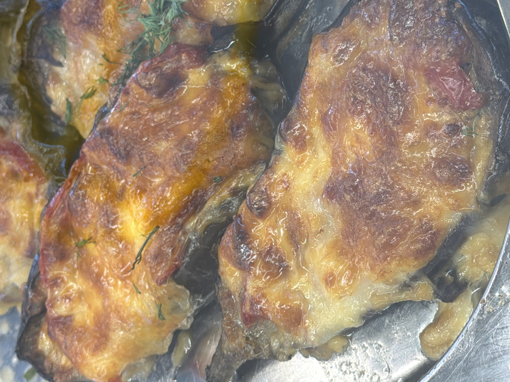 A close-up, overhead view of several portions of baked eggplant are presented in a metallic serving dish. The eggplant is sliced lengthwise and topped with melted cheese that has browned and bubbled from baking. Some of the eggplant skin appears slightly charred. A few sprigs of fresh green herbs, possibly dill or thyme, are visible scattered across the top of the dish. The dish is set on a reflective surface. The image does not contain any information about the city of Sochi, Russia.