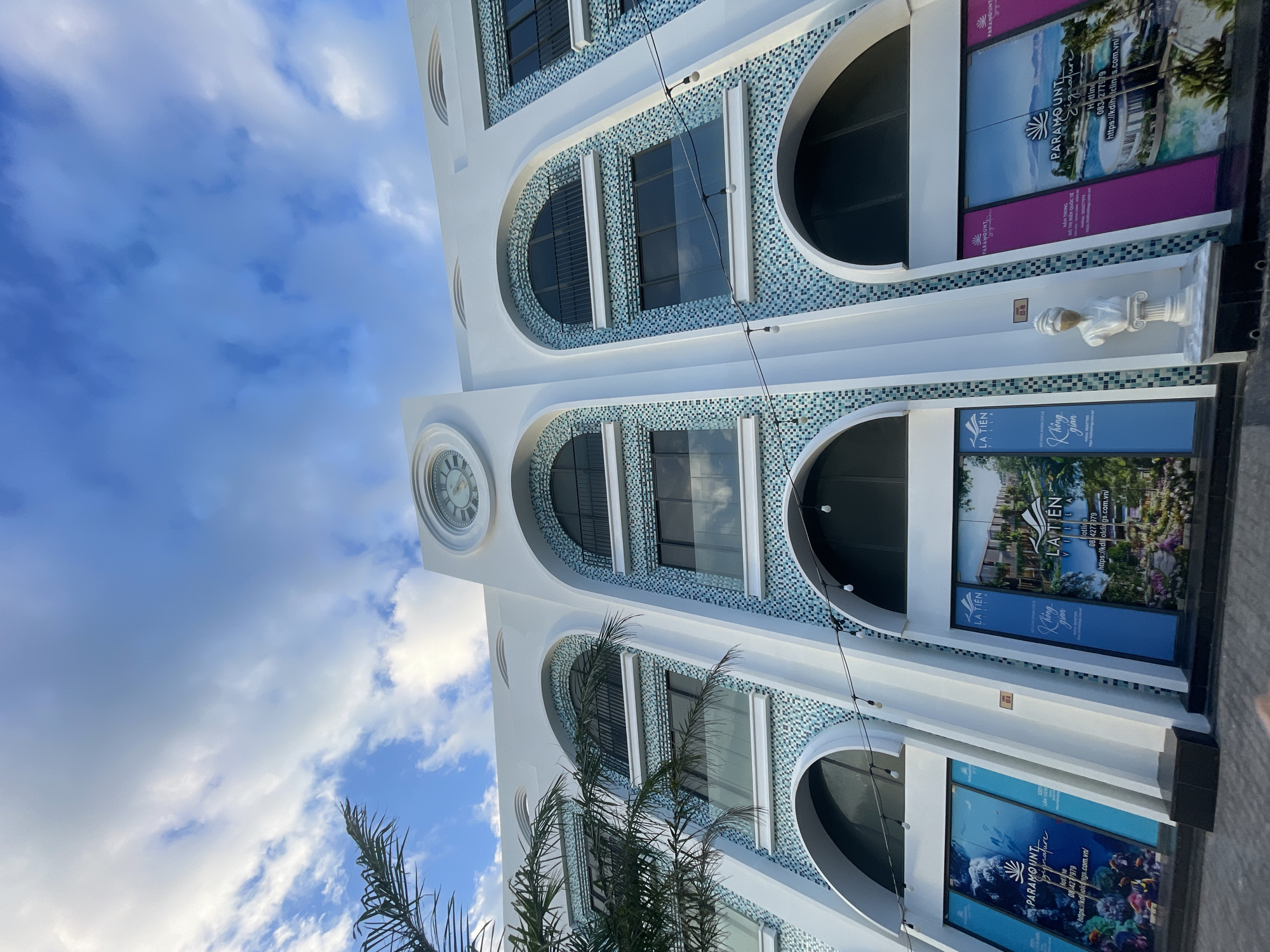 The image shows a low-angle shot of a white building with arched windows and blue mosaic tile accents, set against a partially cloudy sky. The building features a clock on its facade and promotional banners for "LA TIEN VILLA" and "PARAMOUNT Signature", indicating real estate or property advertisements. These banners display phone numbers and websites with a city code indicating they are located in the city of Bac Nha Trang Ward, Vietnam. A palm tree is visible in the lower portion of the frame, adding a tropical element to the scene.
