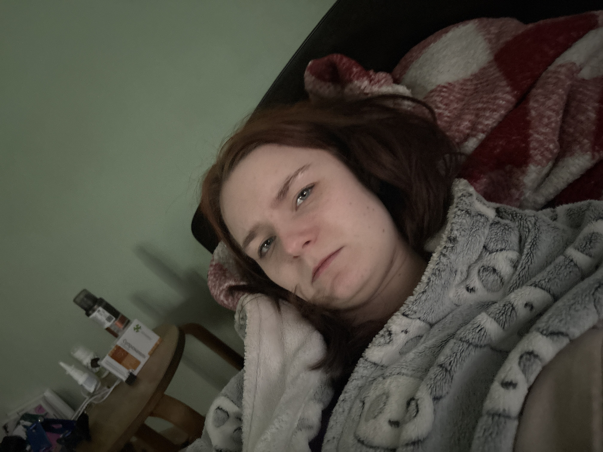 A young adult woman with reddish-brown hair and blue eyes is captured in a close-up, lying down indoors. She is partially covered by a soft, grey, fuzzy blanket or robe adorned with a subtle, possibly animal-themed, pattern. Behind her head and shoulders is a red and white plaid blanket. The woman's expression is neutral, appearing somewhat tired or somber, as she looks directly at the camera. Her face shows some freckles and a slight flush on her cheeks.
The background consists of a light green wall and a dark headboard or pillow. To the left, on a small, light-brown wooden bedside table, are several items. These include various bottles, one with a dropper, and a white spray bottle. A small, light-colored box is also visible, featuring a green cross symbol and partially legible text that appears to read "Ophth..." (likely part of "Ophthalmic") and "Compounds" or "Compositions." The lighting in the room is dim, suggesting it might be evening, night, or a room with subdued illumination. The scene conveys a sense of quiet rest or possibly convalescence. While the image itself offers no specific visual cues to the location, the provided context indicates an unknown location in Russia.
