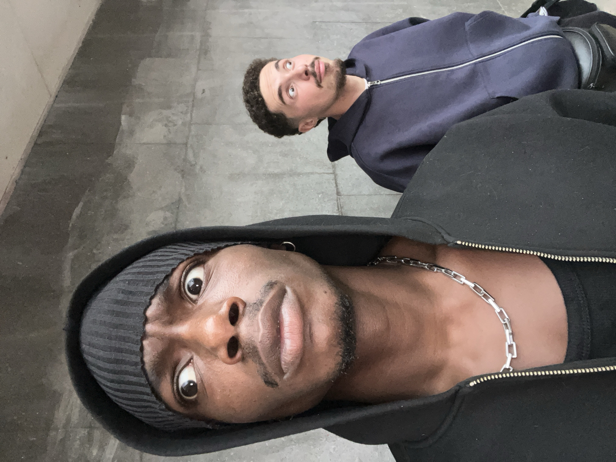 This low-angle photograph captures two men looking upwards with wide, curious expressions, possibly lying down or leaning back on a grey concrete surface in what appears to be an urban public space, consistent with Barcelona, Spain.
In the foreground, a Black man with a short beard is prominent, wearing a dark grey striped beanie and a black zippered hooded jacket. A noticeable silver chain adorns his neck. His eyes are wide open, fixed directly upwards, conveying a sense of surprise or intense observation.
Behind him and slightly to the right, another man with curly brown hair, a mustache, and a short beard is visible. He wears a dark blue zippered hooded jacket. His head is tilted back, and his eyes are also wide open, gazing upwards and to his left, mirroring the foreground subject's focused attention. A dark bag or object is partially seen behind him.
The background consists of a textured, grey concrete or tiled floor, possibly part of an underpass, subway station, or a sheltered public area. A light-colored wall or vertical structure is visible on the far left edge of the frame. The lighting is diffused, suggesting an indoor setting or an overcast day. No text is visible in the image. Both individuals seem captivated by something unseen above them, creating an engaging and mysterious interaction with their environment.