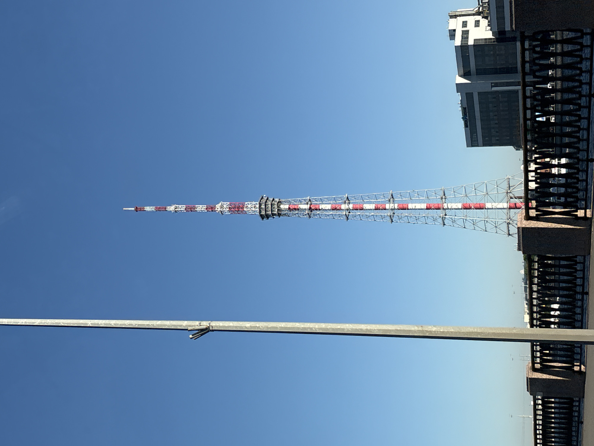 The image shows a tall, slender, red and white striped TV tower in Saint Petersburg, Russia. The tower is made of a lattice framework, and it extends high into a clear blue sky. A lamppost stands in the foreground. A building is partially visible behind the tower, to the right, and a wrought-iron fence is also present to the right, close to the edge of the frame.