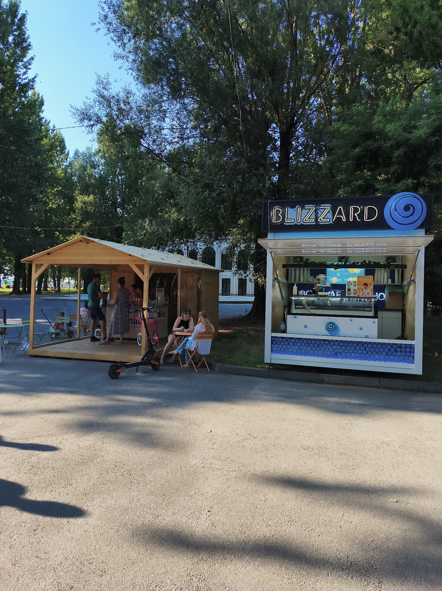 This is an outdoor scene on a sunny day, likely in the city of Ternopil, Ukraine, featuring two food stands. On the right is a modern, brightly lit stand with the prominent neon sign "BLIZZARD" and a blue swirling logo, advertising ice cream or similar frozen treats. The stand is open at the front, with a glass display case showing frozen products and a person behind the counter. Below the counter, a decorative element with blue spheres and lights is visible. On the left is a simpler, wooden, open-air kiosk with a sloped roof. Several people are present at this kiosk: a man in a green shirt stands and appears to be interacting with a woman in a patterned dress. Two young women are seated at small tables outside the kiosk, one facing the camera and the other with her back to it. An electric scooter is parked in front of the wooden kiosk. The ground is paved with asphalt, and shadows from trees are cast across it. In the background, there are lush green trees and a building with arched windows.