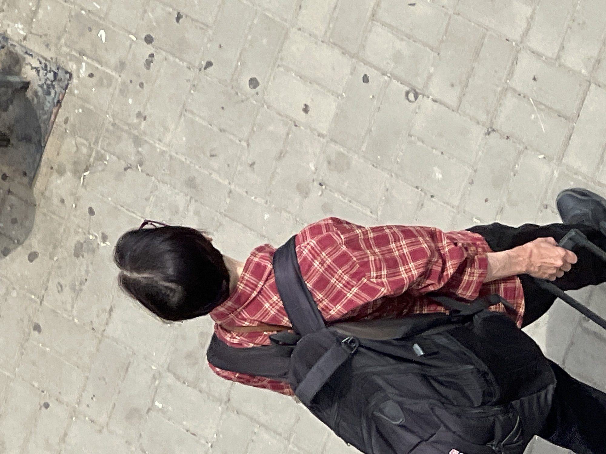 The image shows a person from a high-angle perspective, walking on a paved walkway. The person is wearing a red and white plaid shirt, a dark backpack, and dark pants. They appear to be holding onto the handle of a rolling suitcase, which is mostly out of frame to the right. The ground is made of light-colored rectangular paving stones, with some scattered debris and dark spots, possibly from rain or dirt. The lighting suggests it is daytime, and the weather is overcast or the light is diffused. The scene appears to be an urban environment, likely a street or park. The person's back is mostly to the camera, so their facial expression is not visible, but their posture suggests movement and purpose. No text is visible in the image.