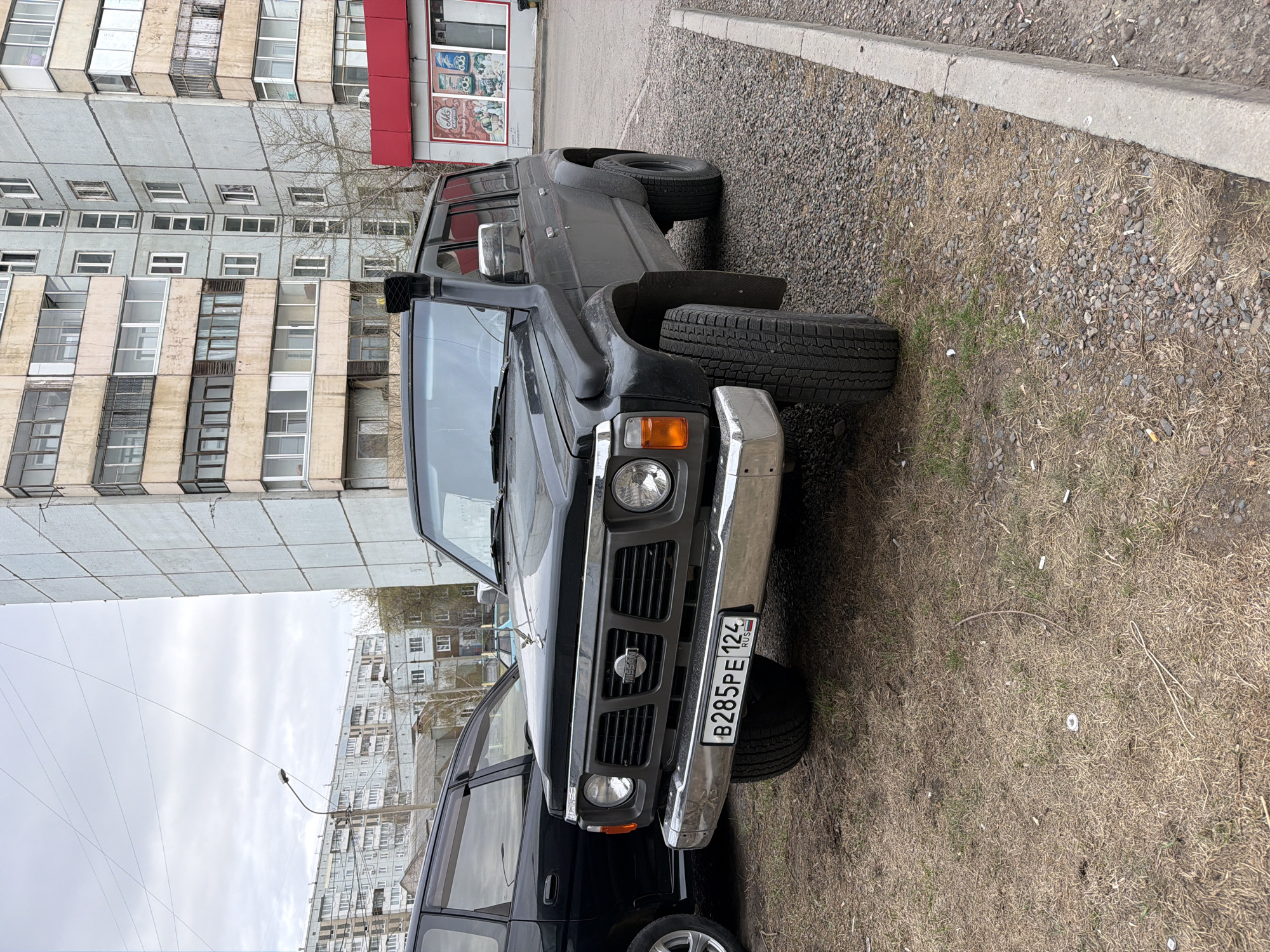 The image shows a black Nissan Patrol SUV parked in what appears to be a semi-urban setting in Krasnoyarsk, Russia, indicated by the license plate "B285PE 124 RUS". The vehicle is positioned alongside a curb and next to a building with multiple floors and windows. In the background, another building with a red awning is visible. The ground surface consists of a mix of gravel, grass, and discarded materials, including what appear to be cigarette butts. There are no people visible in the image.