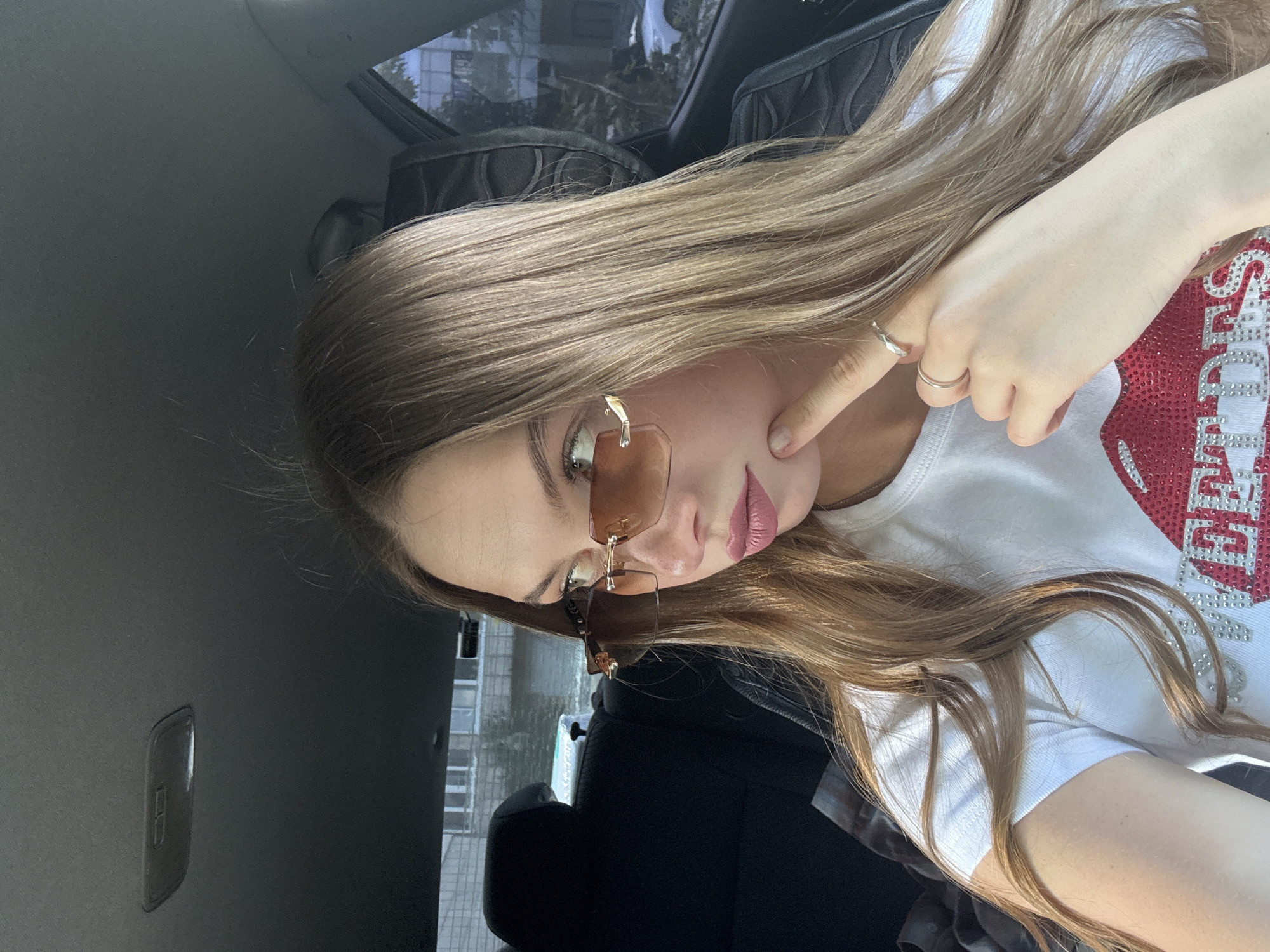 A young woman with long, blonde hair, wearing sunglasses and a white t-shirt with red and silver embellishments, poses for a selfie inside a car. She is touching her cheek with her index finger. The interior of the car appears to be dark, with black upholstery. Outside the car window, a brick building and trees are visible, suggesting an urban or suburban setting. The lighting suggests it is daytime. The woman's expression is playful or suggestive, with her lips slightly parted. The embellishments on her t-shirt appear to form text or a design, but are not clearly legible. The overall mood is casual and self-assured. The image is taken from a low angle, looking up at the woman.