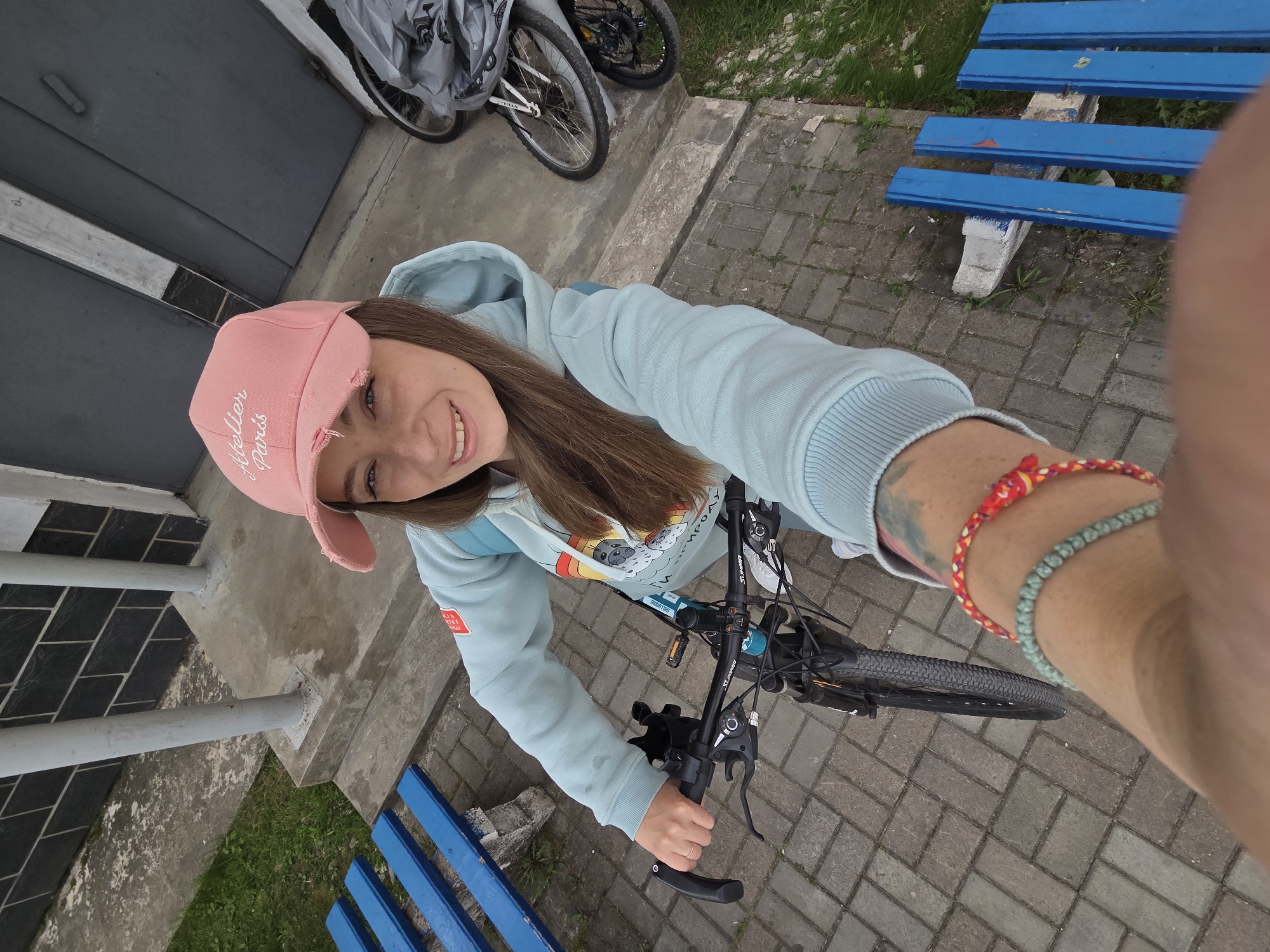 A young woman is taking a selfie while standing over a bicycle. She is wearing a light blue hooded sweatshirt and a pink cap with white lettering. She has long brown hair and is smiling at the camera. On her wrist, she wears two bracelets, one red and one green. She is holding the handlebars of a black bicycle. In the background, there are two bicycles covered with gray tarps, a gray building, and blue benches. The ground is paved with gray bricks. The location appears to be in a city, specifically Birobidzhan, Russia.