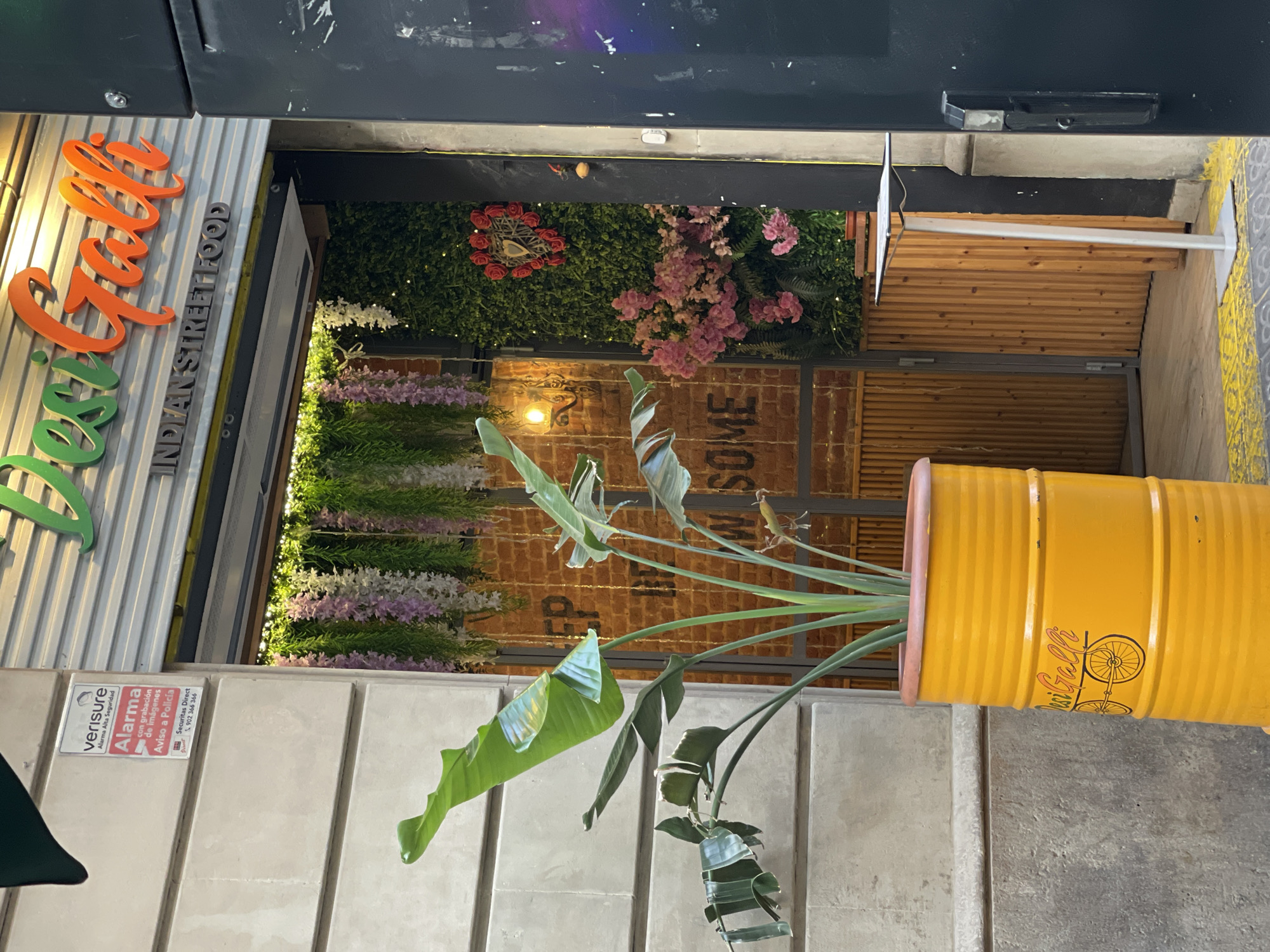 The image shows the entrance to "Desi Galli Indian Street Food" in Barcelona, Spain. The restaurant has a sign with the name in large orange and green letters, and below it, "INDIAN STREET FOOD" in grey lettering. The entrance is decorated with artificial greenery, hanging purple and white flowers, and a heart-shaped wreath made of red roses and twigs. To the left of the entrance, a large yellow planter shaped like a barrel contains a tall leafy plant. A "Verisure Alarma" sticker is visible on a building pillar to the left of the planter. A thin, silver stand with a small square top is placed to the right of the entrance. The entrance itself is dark and leads into the restaurant.