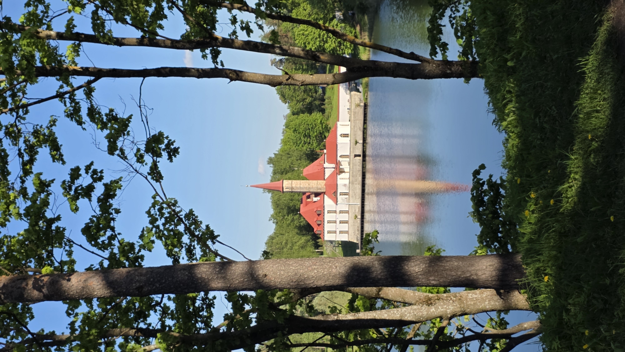 The image shows the Priory Palace in Gatchina, Russia, viewed through tree branches. The palace, with its red roof and light-colored walls, is reflected in the water. Trees and foliage are visible on the banks of the water. The sky is blue.
