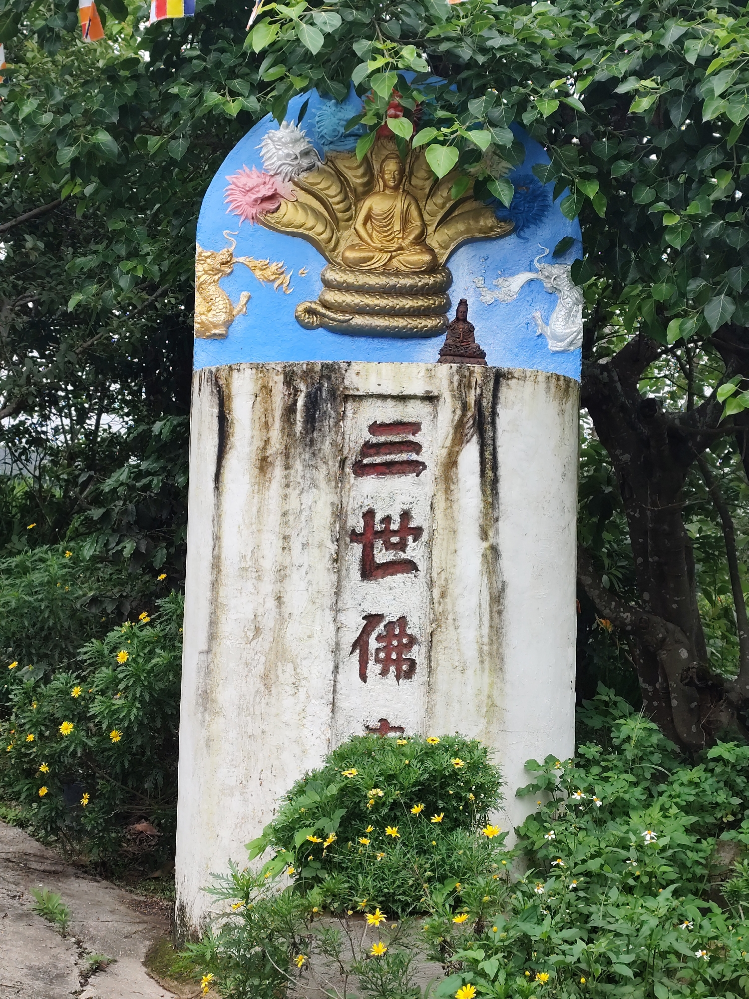 A weathered, cylindrical monument stands prominently amidst lush green foliage and yellow flowering bushes, characteristic of an outdoor setting like Phường Xuân Hương - Đà Lạt, Vietnam. The pillar's main body is off-white, heavily stained with vertical streaks of dark grime or mold, indicating age and exposure to the elements.
Bold red Chinese characters are vertically inscribed on the front of the pillar, reading "三世佛" (Sān Shì Fó), which translates to "Buddhas of the Three Ages" or "Three World Buddhas."
The upper, arched section of the monument features a vibrant blue background. Centrally, a golden Buddha figure is depicted in a meditative pose, seated upon a coiled naga whose multi-headed hood shelters him. To the left of the Buddha, a golden, dragon-like creature appears to exhale fiery breath or mist, accompanied by a pink, stylized floral element. On the right, a white dragon-like figure mirrors the golden one, with a white floral or cloud-like motif above it. A small, dark brown Buddha statue is placed at the base of the central golden Buddha relief.
Overhead, colorful prayer flags in traditional Buddhist hues of red, blue, yellow, white, and green are strung amongst the dense, large-leafed trees, likely Bodhi trees, which partially shade the monument. The base of the monument is surrounded by flourishing green bushes dotted with small yellow flowers, and a narrow, cracked path curves around its left side. The scene, captured during daytime, conveys a serene and sacred atmosphere.
