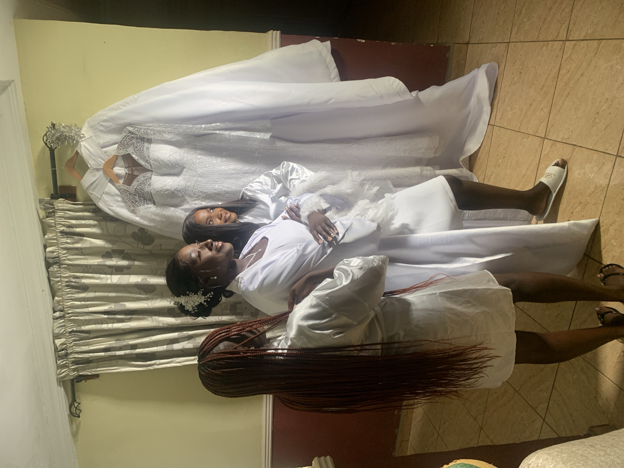The image shows two Black women lying on top of what appears to be wedding dresses. One woman wears a white robe, has dark hair pulled back with white floral accents, and is smiling. The other woman has long brown braids and wears a white robe with a feathered collar. A wedding dress hangs on a hanger in the background. The location is indoors with tiled flooring, a floral-patterned curtain and light-colored walls. The city of Barikin Sabon Gidan Kanat, Nigeria is not discernible in the image.