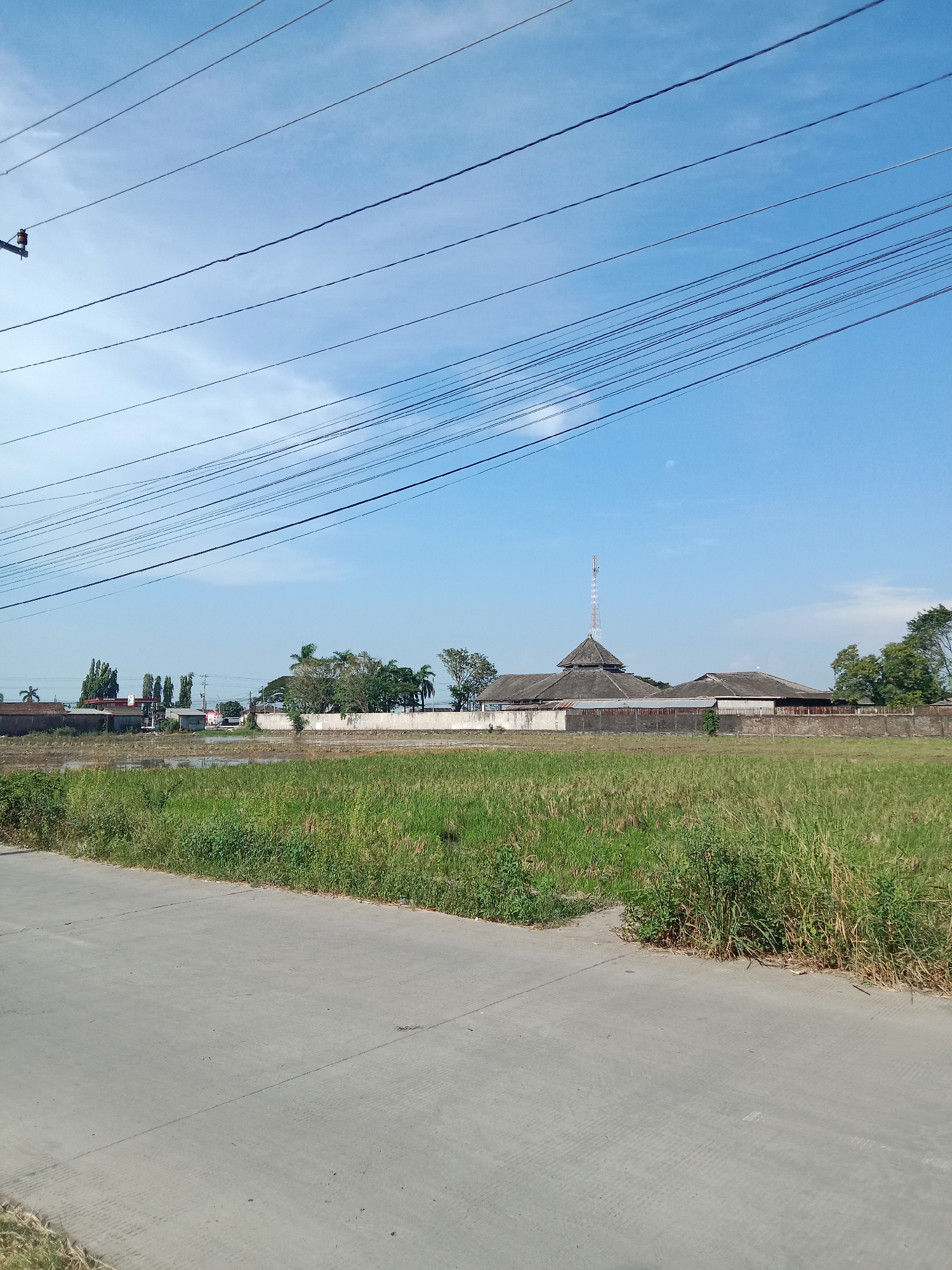 The image shows a landscape in Cangkring, Indonesia, under a clear, blue sky. Overhead are a network of power lines. In the foreground is a gray concrete road with grassy verges. Beyond the road, there's a field of tall, green grass and weeds. In the distance, a low wall surrounds a building complex featuring a multi-tiered roof structure, topped by a radio or telecommunications tower. Additional buildings are visible adjacent to the main structure. Trees are interspersed around the buildings. In the far background, there is a hint of other structures and trees.