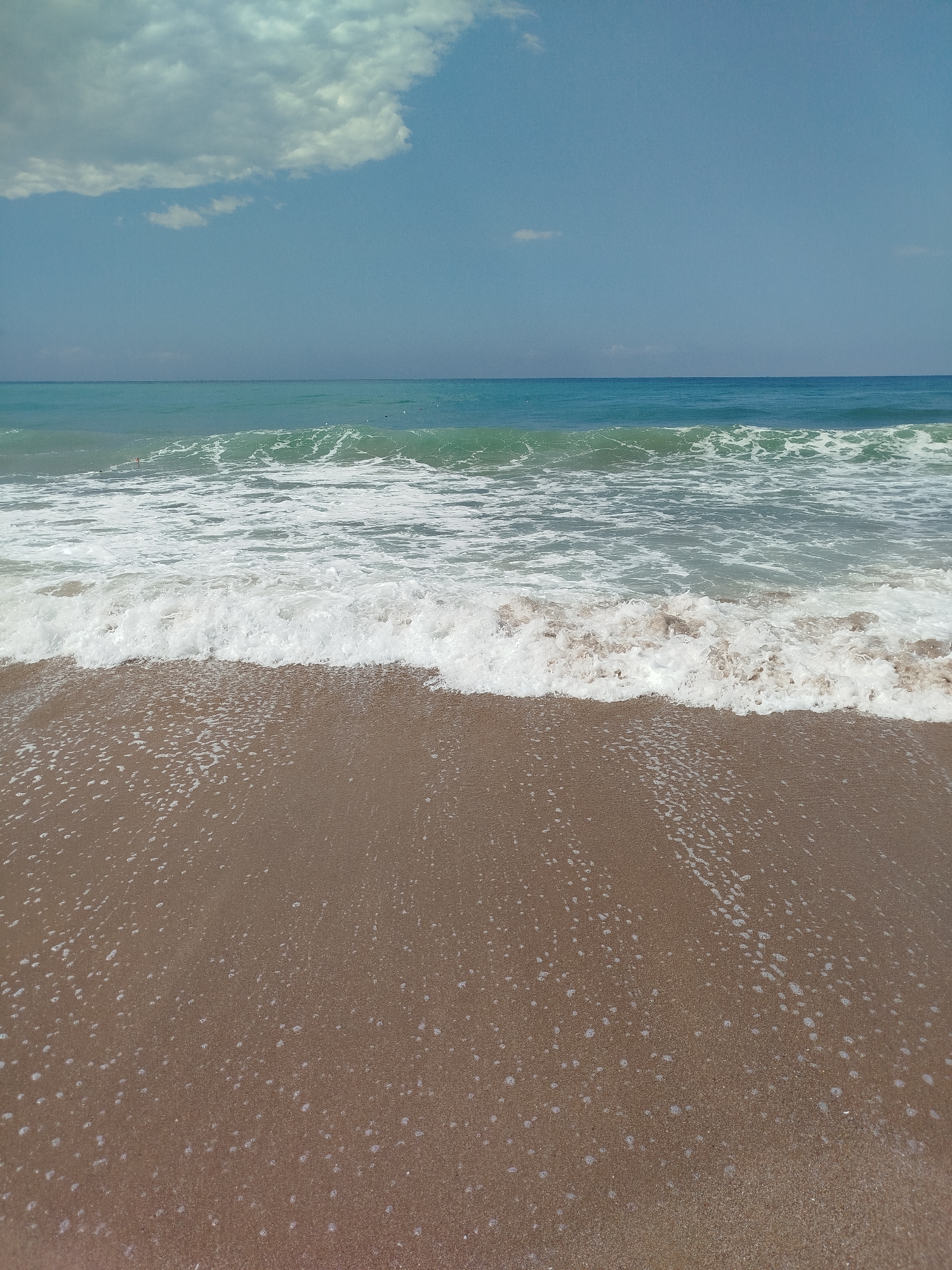 A tranquil scene of a sandy beach meets the turquoise waters of the Mediterranean Sea under a clear blue sky. Gentle waves are rolling onto the shore, leaving a foamy white trail on the wet sand. A large, fluffy cloud drifts lazily in the upper left corner of the frame. The horizon is sharp, separating the sea from the sky. The sand is a warm brown color, dotted with small, light-colored pebbles and scattered shells. The water is a vibrant shade of blue-green, with white caps on the breaking waves. The overall atmosphere is one of peace and natural beauty, characteristic of the coastline in Manavgat, Turkey.