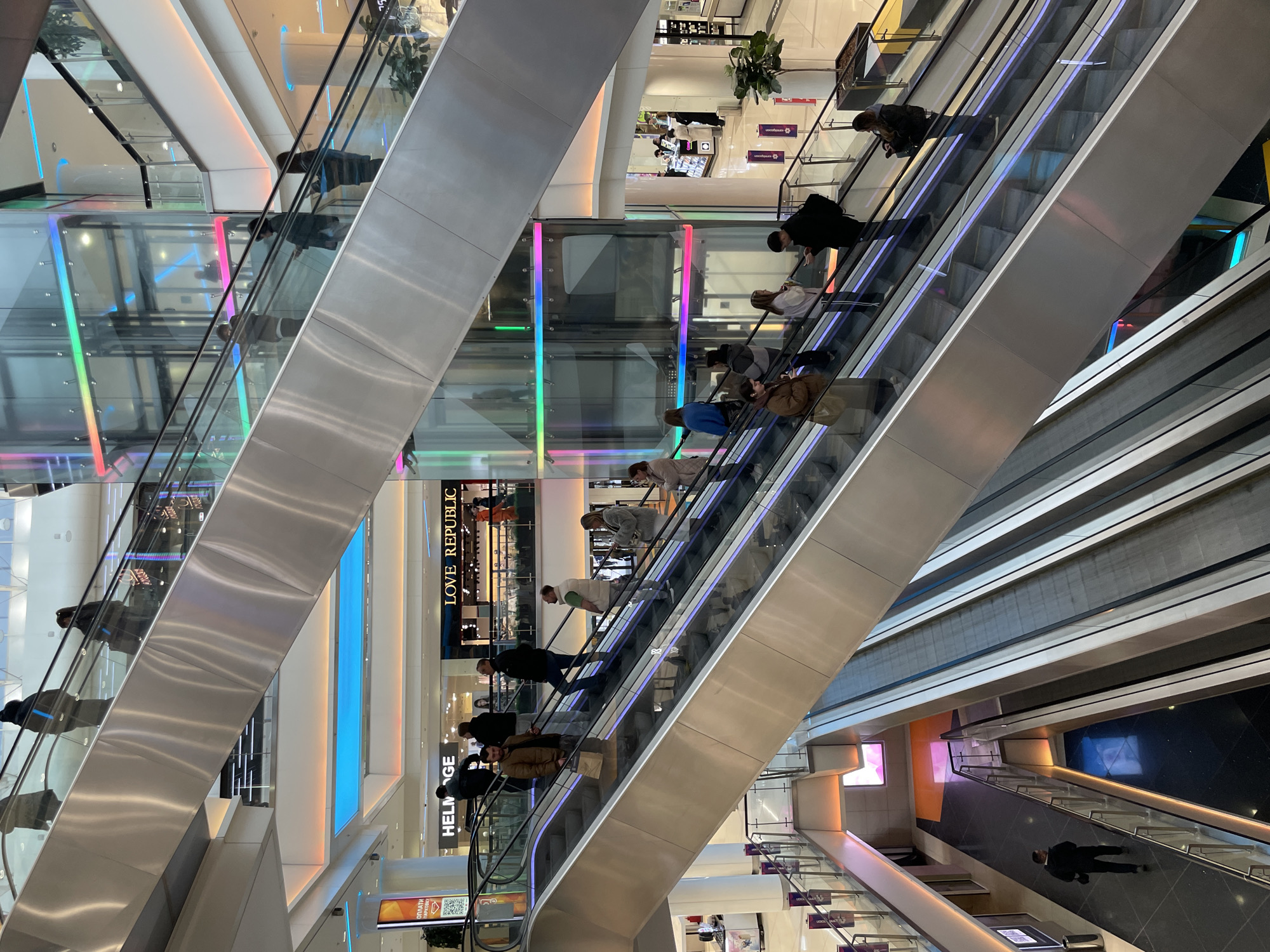 The image depicts the interior of a multi-story shopping mall, possibly in the city of Unknown, Unknown. The focal point is a series of escalators that intersect at various levels. People are riding the escalators in both upward and downward directions. The mall's interior has a modern design with glass railings on the escalators and multiple levels visible, each with different stores and lighting features. Some stores display signage, such as "LOVE REPUBLIC" and "HELIDGE." Colorful LED lighting is integrated into the architecture, enhancing the modern aesthetic. The overall environment suggests a bustling retail space.