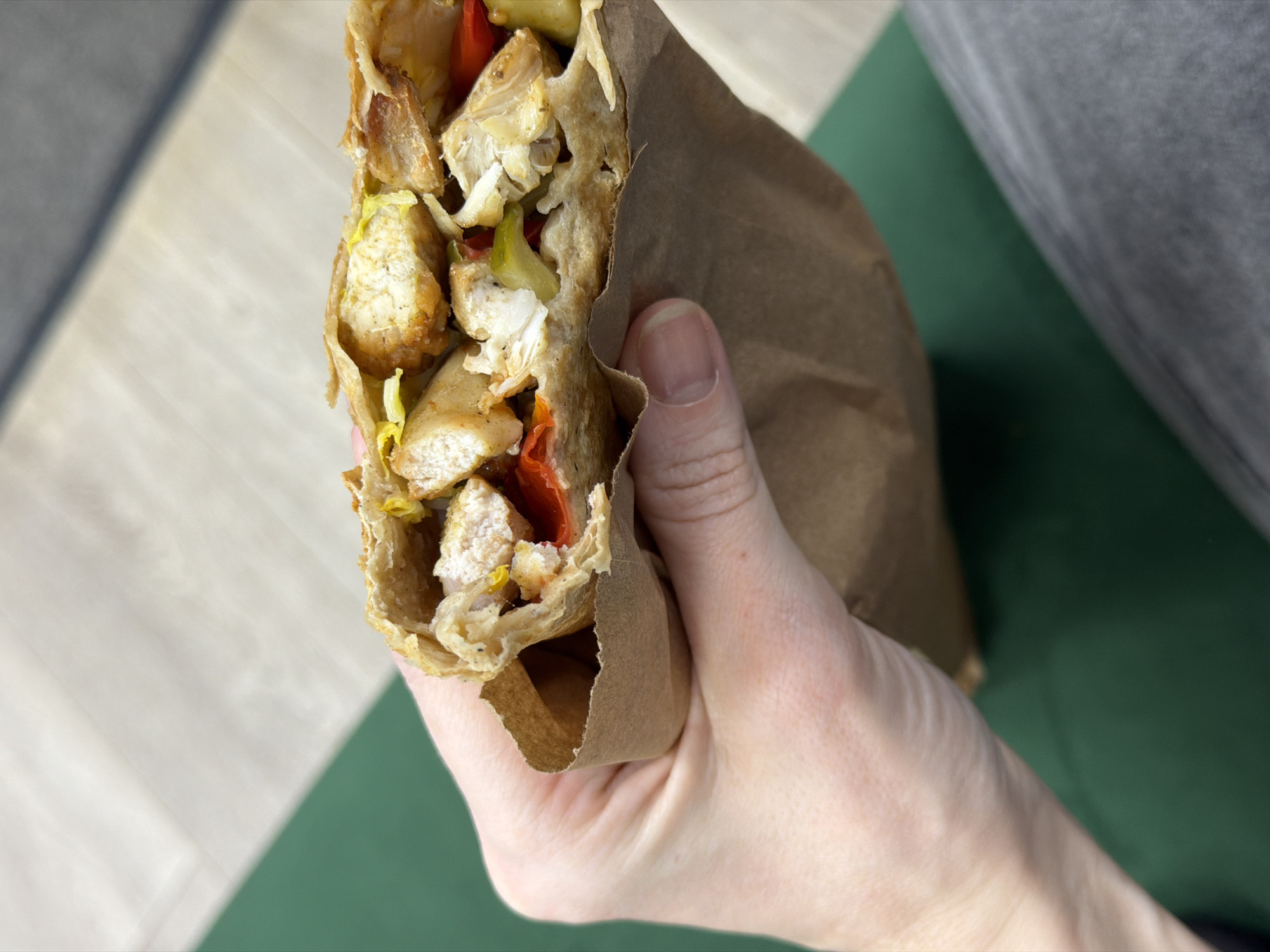 A hand holds a partially eaten gyro in a brown paper wrapper. The gyro appears to contain chicken, red peppers, and lettuce. The background consists of a multi-toned green surface meeting a light-colored surface, with a portion of a grey garment visible. The location is likely indoors. There is no evidence in the image to place it in Perm, Russia.
