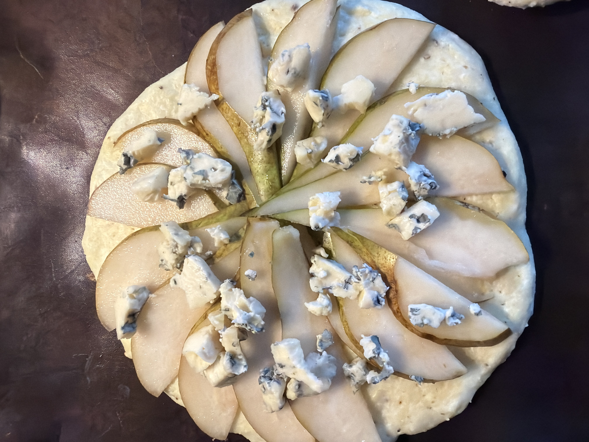 A top-down, close-up shot shows a partially prepared food item resembling a pizza or flatbread. The base appears to be a light-colored dough. Thinly sliced pear segments are arranged in a radial pattern across the surface, with chunks of blue cheese scattered over the pears. The composition sits on a dark brown surface. The scene contains no information about Saint Petersburg, Russia.