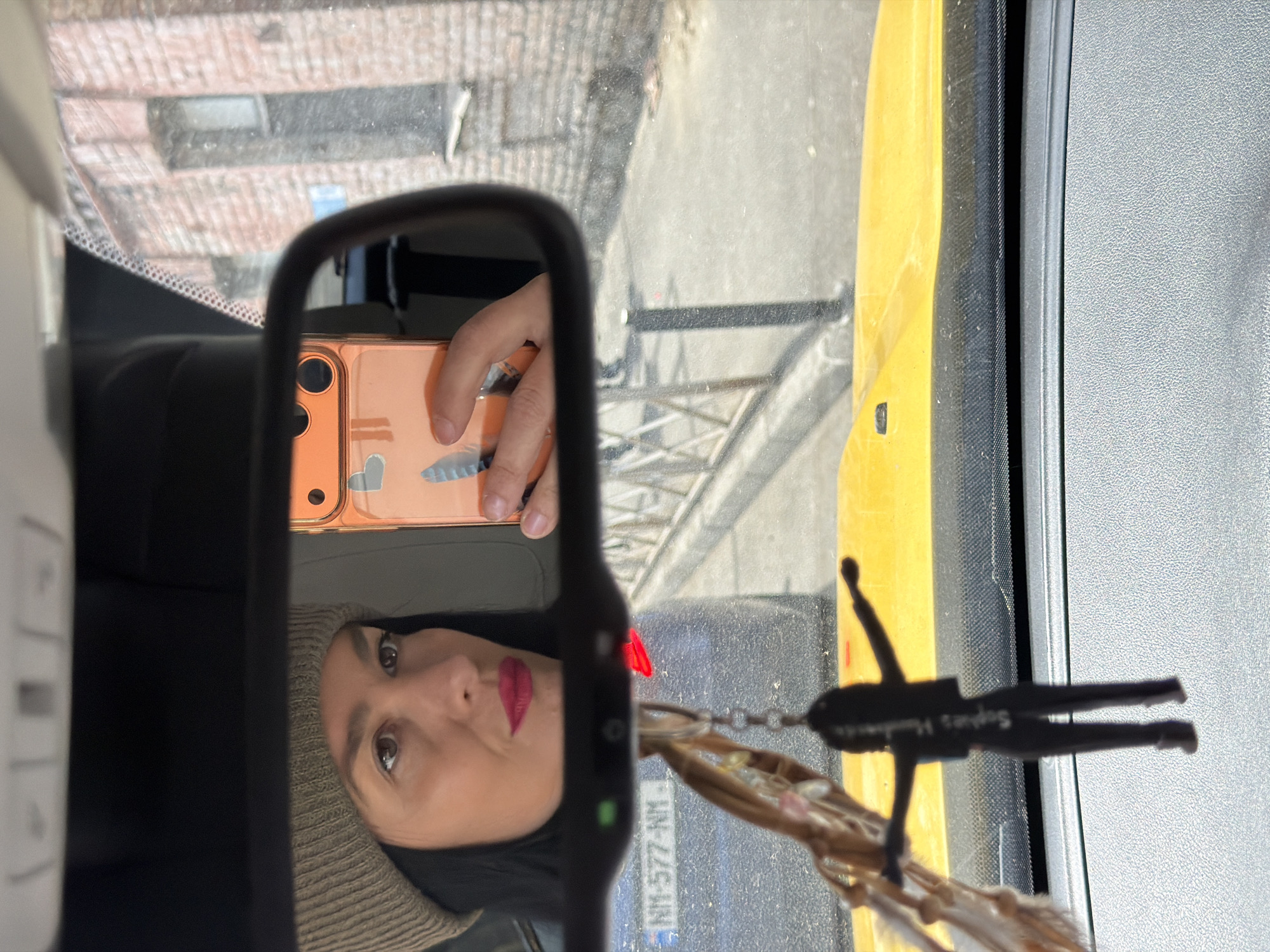 This media file captures a selfie taken from inside a car in Tbilisi, Georgia, with the subject's reflection visible in the rearview mirror. The image features a woman, likely the driver, wearing a light brown knit beanie. She has dark hair and red lipstick, and is looking towards her smartphone, which she holds up. Her hand and the phone, encased in a light orange/pink case with some faint white/blue designs, are reflected in the mirror.
Hanging from the rearview mirror is a black, stylized figure or air freshener with the text "Sopho's Handmade" visible on it.
Outside the car, visible through the windshield and reflected in the mirror, is a bright daytime scene. A large yellow vehicle, possibly construction equipment or a truck, is prominent on the right side. In the background, a brick building is visible. A license plate from another vehicle, partially visible in the reflection, reads "NM-5777-NM", consistent with Georgian license plate formats, confirming the location as Tbilisi, Georgia. The weather appears clear and sunny. The overall impression is a casual, everyday moment captured inside a vehicle.