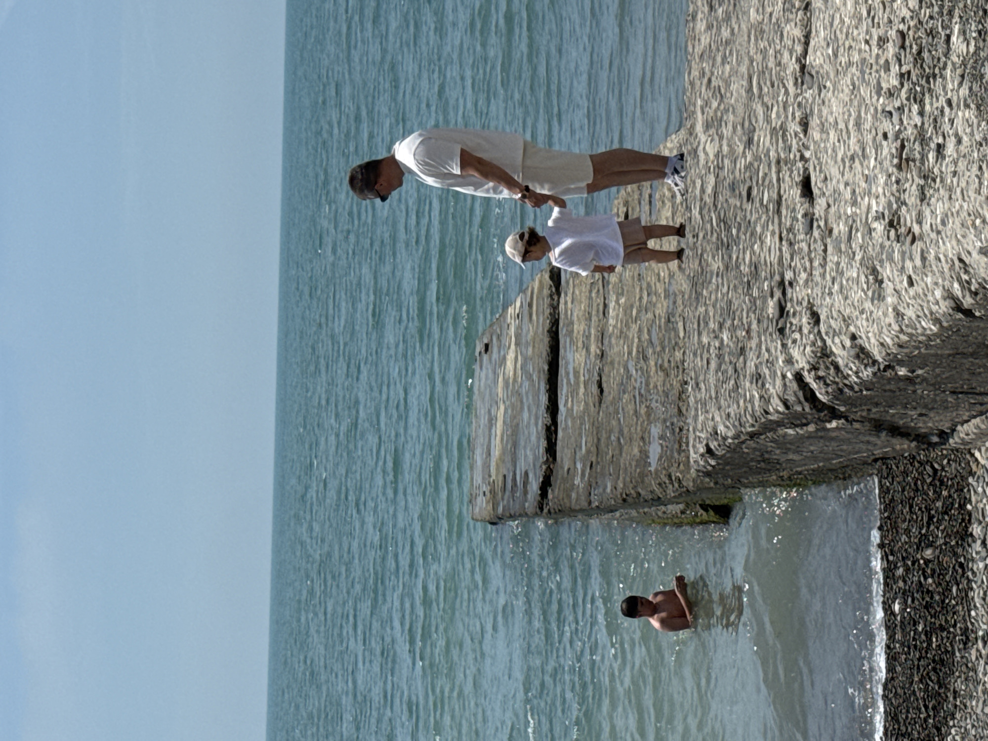 The image depicts a bright, sunny coastal scene in Sochi, Russia, featuring a concrete pier extending into the sea. The calm, blue-green water of the sea meets a clear, light blue sky at the horizon.
On the textured, grey concrete pier, a man and a young child stand. The man, appearing middle-aged with short dark hair and sunglasses perched on his head, wears a white short-sleeved t-shirt, light-colored shorts, and white athletic shoes with dark soles. He stands upright, facing slightly left, looking downwards, and holds the right hand of the child. The child, appearing to be a toddler or preschooler, wears a light-colored baseball cap, a white t-shirt, and tan shorts. The child stands facing away from the camera, looking towards the water.
To the left of the pier, in the shallow water, a third individual, appearing to be an adolescent or young adult, is partially submerged to the chest. This person is shirtless and looks down at an object held in their hands. The water around them shows slight ripples. The immediate foreground at the bottom left of the image reveals a small section of a pebble beach where the water meets the shore.