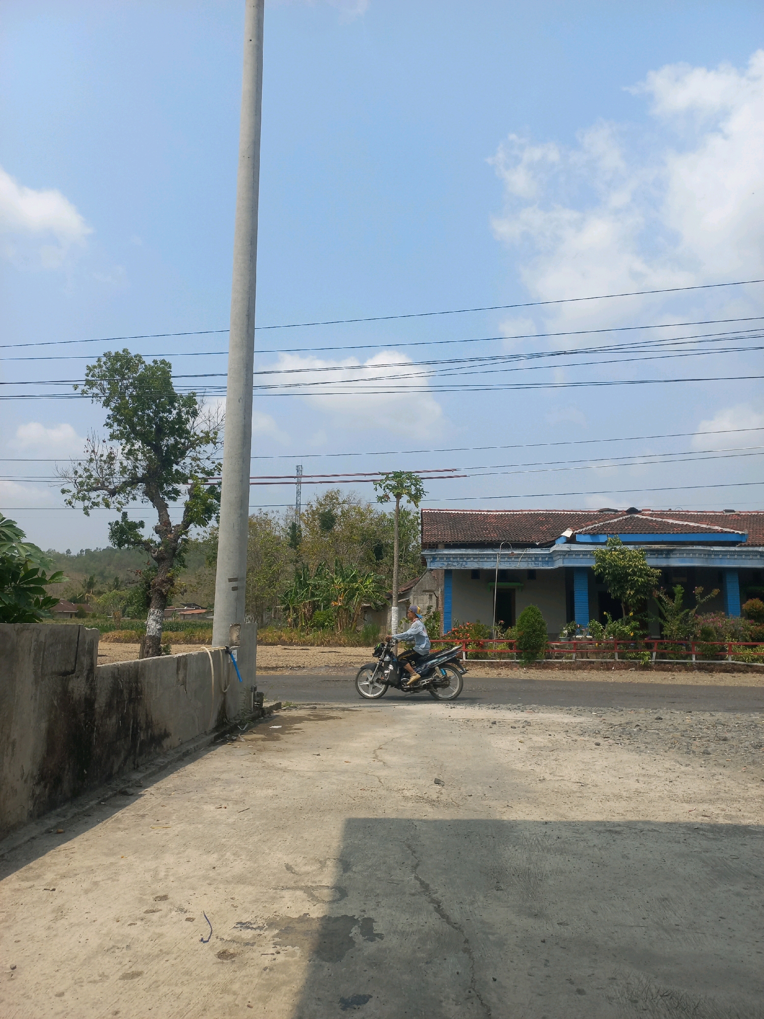A person is riding a motorcycle along a paved road in Baturetno, Indonesia. The rider is wearing a blue long-sleeved shirt and a light-colored headscarf. The motorcycle is dark in color with silver accents. A light-colored concrete wall is in the foreground to the left, casting a shadow. Behind the wall, a large tree with sparse green leaves stands beside a utility pole. The road runs parallel to a house with a blue-trimmed roof and a red fence. Various trees and vegetation are visible in the background, with a hill in the distance. The sky is blue with scattered white clouds. Electrical wires are strung across the upper portion of the image.