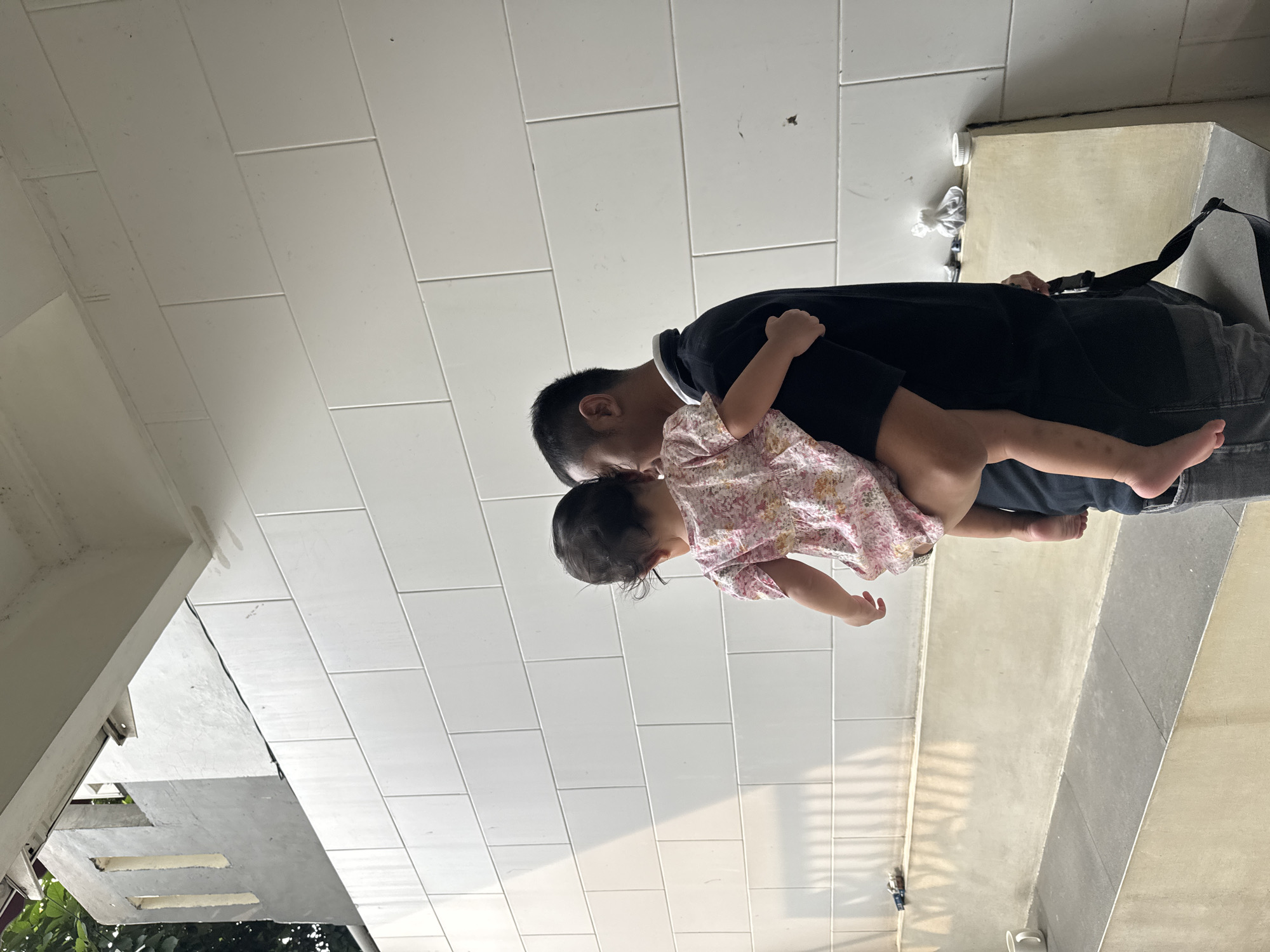 The image shows a man carrying a young child. The man has short black hair and is wearing a black shirt and gray jeans. The child is wearing a floral print dress. The background consists of white wall paneling, concrete surfaces, and some visible foliage. The location is the city of Bekasi, Indonesia.