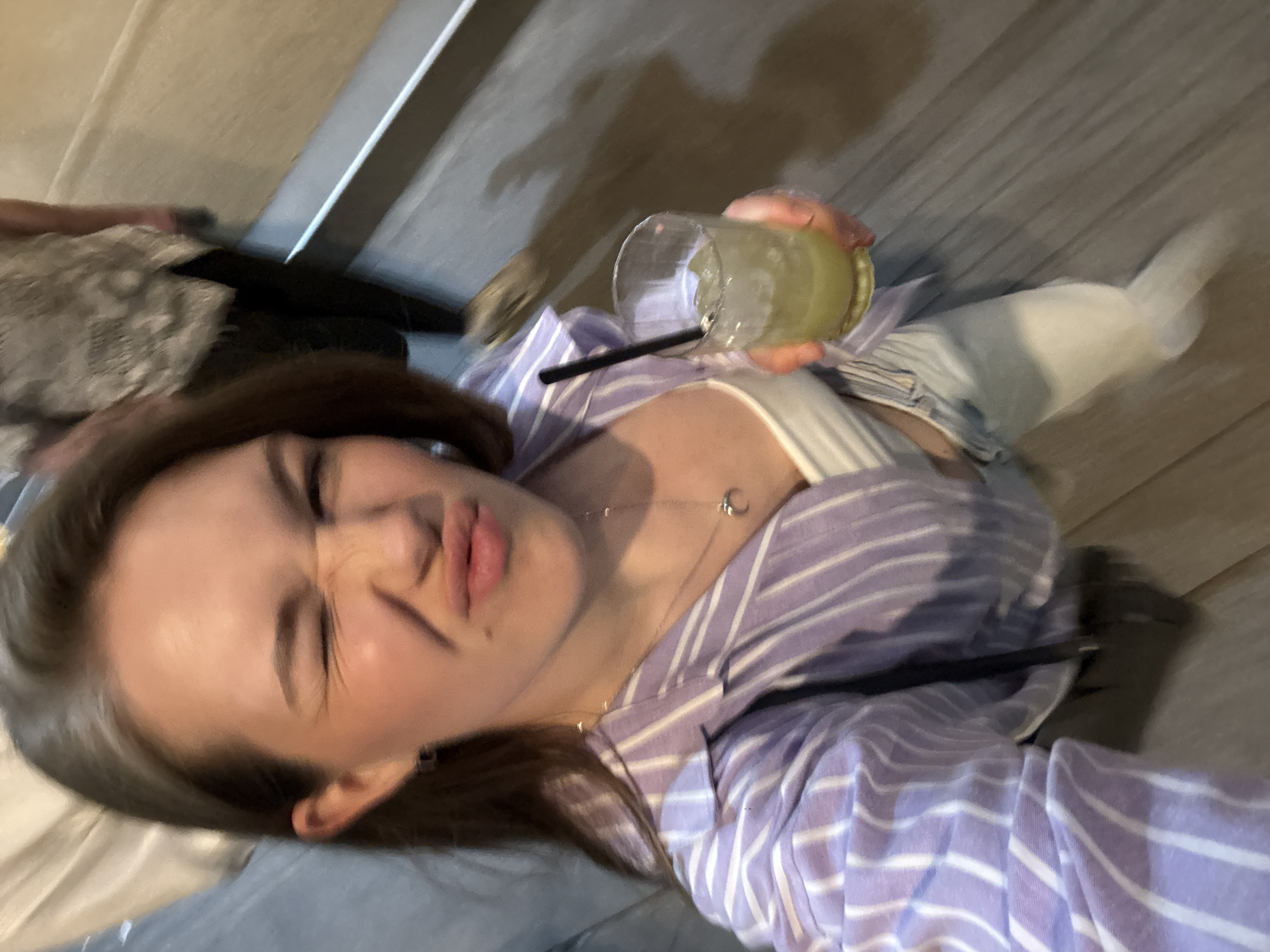 A young woman is taking a selfie. She has light skin, brown hair with graying roots, and is making a scrunched-up, playful face. She is wearing a striped lavender and white shirt with a white tank top underneath. A silver necklace with a pendant is visible. She is holding a glass with a light-colored beverage and ice. A black straw is in the glass. The location is likely indoors, possibly in a bar or restaurant based on the style of drink. The background is blurry, but it appears to be a wall and part of a table or seating area. There is no visible evidence to identify Belgrade, Serbia as the location.