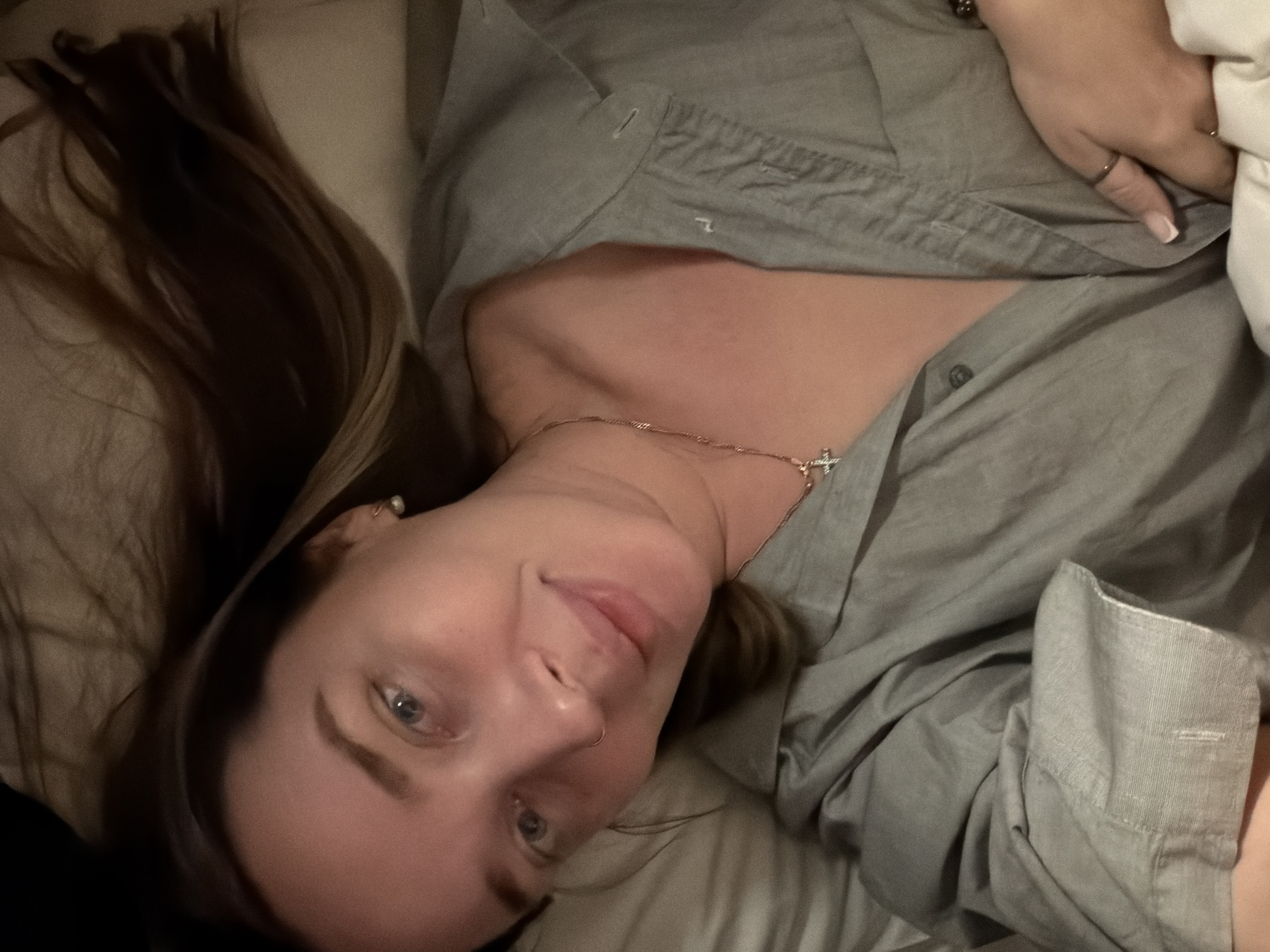A young woman with long, dark blonde or light brown hair lies on light-colored bedding, looking directly at the camera with a gentle, soft smile. Her head is slightly tilted, and her light-colored eyes (blue or grey) are open and engaging. She has fair skin and a small stud nose piercing.
She is wearing a casual, light grey button-up shirt, which appears somewhat rumpled, with the top buttons undone, revealing a delicate rose gold or silver chain necklace with a small cross pendant. A small, pearl-like stud earring is visible in her left ear, and a thin ring adorns the ring finger of her right hand, which rests near her chest, partially obscured by bedding. Her nails are neatly manicured with a light tip.
The setting appears to be indoors, likely a bedroom, characterized by soft bedding and pillows that frame her head and shoulders. The lighting is dim and warm, suggesting an evening or nighttime atmosphere, creating a relaxed and intimate mood. There are no discernible location-specific cues within the image, though the provided context indicates the location is Moscow, Russia. An indistinct embroidered symbol can be seen on her shirt. No visible text is present.