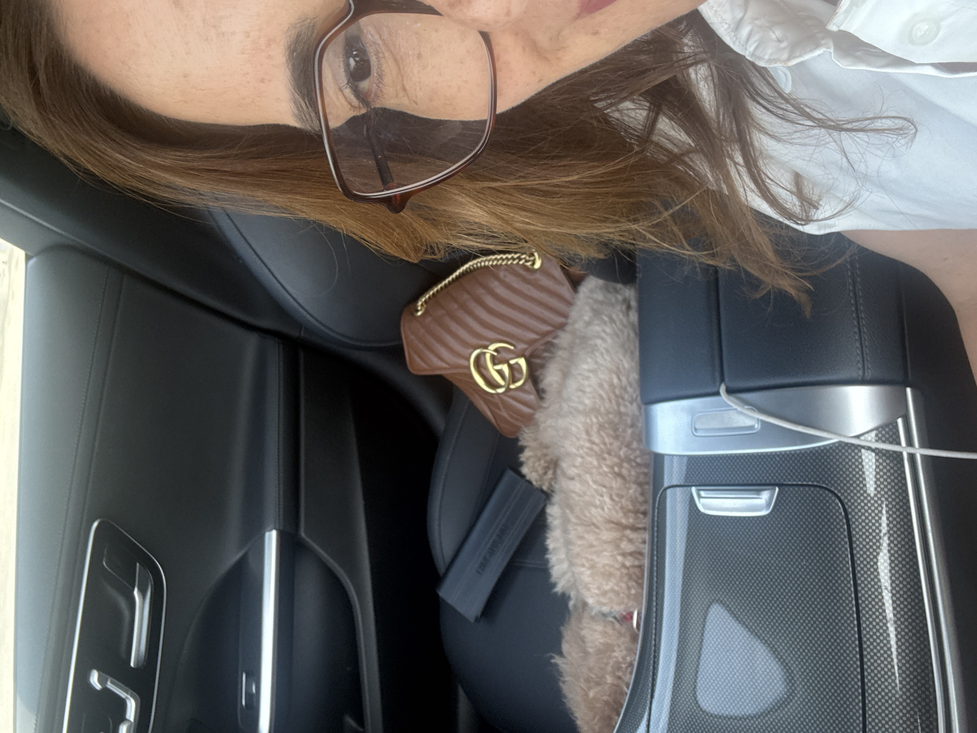 The image provides an interior view of a modern car, likely taken in Alcobaça, Portugal. The main subject is a person whose face is partially visible on the left side of the frame. They have medium brown hair and are wearing brown-framed glasses, with one eye and eyebrow clearly visible. A white collared shirt is seen on their upper body.
In the car's interior, a small, brown, quilted Gucci bag with a gold chain strap and a prominent gold interlocking "GG" logo is resting on a black leather car seat. Beside the bag, a fluffy, light brown object, possibly a small dog or a fur accessory, occupies part of the seat. A black rectangular item, which could be a phone or a small case, is also visible on the seat.
The car's design features black leather upholstery, sleek silver trim accents, and a distinctive carbon fiber pattern on the center console and door panels, indicating a modern and luxurious vehicle. A white charging cable is draped across the center console. The lighting suggests it is daytime. The image appears to be a self-portrait or a casual photo taken from within the car, presented with a 90-degree rotation.