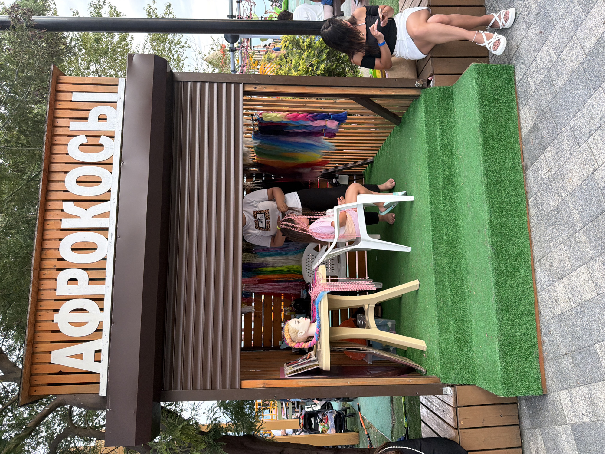 This is a vertical, eye-level shot of a small kiosk selling hair extensions, likely in an outdoor market or amusement park setting. The kiosk has a brown corrugated metal roof and sides, and a wooden facade with large white letters spelling out "АФРОКОСЫ," which translates to "Afro Braids" or "Afro Kosa." This suggests the business specializes in braiding African-style hairstyles.
In front of the kiosk, the ground is covered in green artificial turf, arranged in tiered steps. A mannequin head with brightly colored braids is displayed on a small table. Further inside the kiosk, colorful bundles of hair extensions are hanging from a rack.
Two individuals are visible in the scene. One person, wearing a white t-shirt with a leopard print design and black pants, appears to be an employee or vendor, possibly working on hair. Another person, wearing a pink top and black leggings, is seated on a white plastic chair inside the kiosk.
In the background, through an opening in the kiosk, a playground structure with colorful equipment is visible, suggesting a family-friendly or recreational location. Another person, wearing a black top and white shorts with white sandals, is seated on a wooden bench outside the kiosk, facing away from the camera. The lighting suggests it is daytime, possibly late afternoon or early evening, with overcast conditions. The overall atmosphere seems relaxed and casual. The location cues, such as the playground and the style of the kiosk, point towards a tourist area or a lively public space in Sochi, Russia.