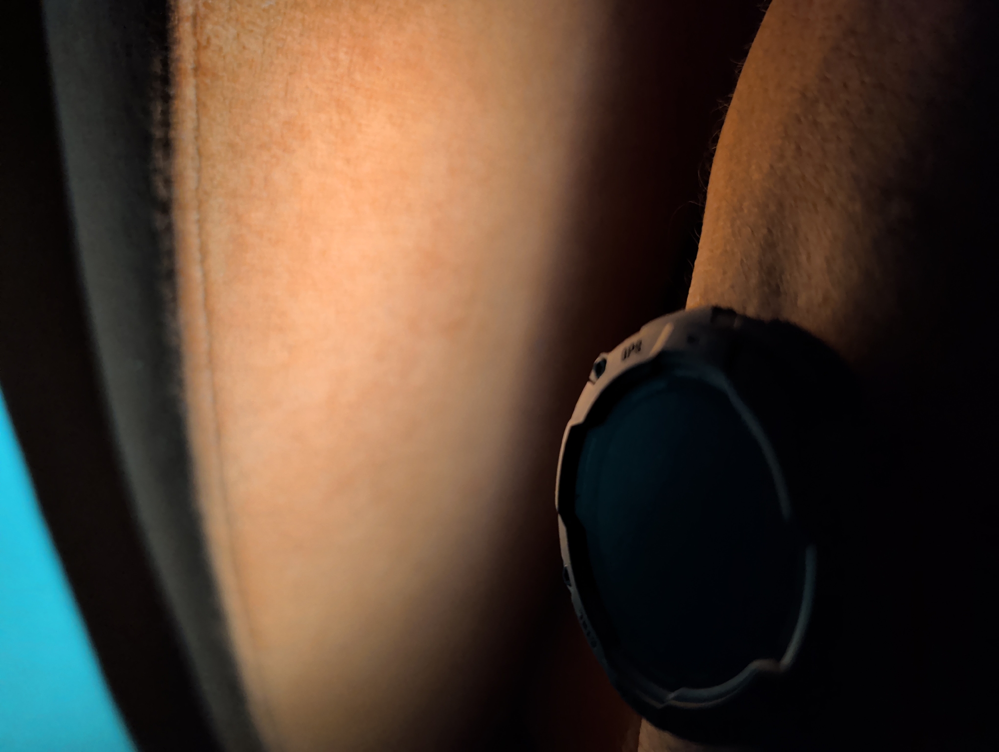 The image shows a close-up of a person's arm with a black digital watch on the wrist. The skin tone is light to medium. The background is a blurred gradient of colors, with a bright teal at the top, transitioning to black, and then to a warm orange-brown tone. The lighting is dramatic, creating strong shadows and highlights, particularly on the arm and watch. There are no discernible elements that place the image in Bishkek City, Kyrgyzstan or any other specific urban location. The scene is devoid of any environmental context beyond the immediate subject and background colors.
