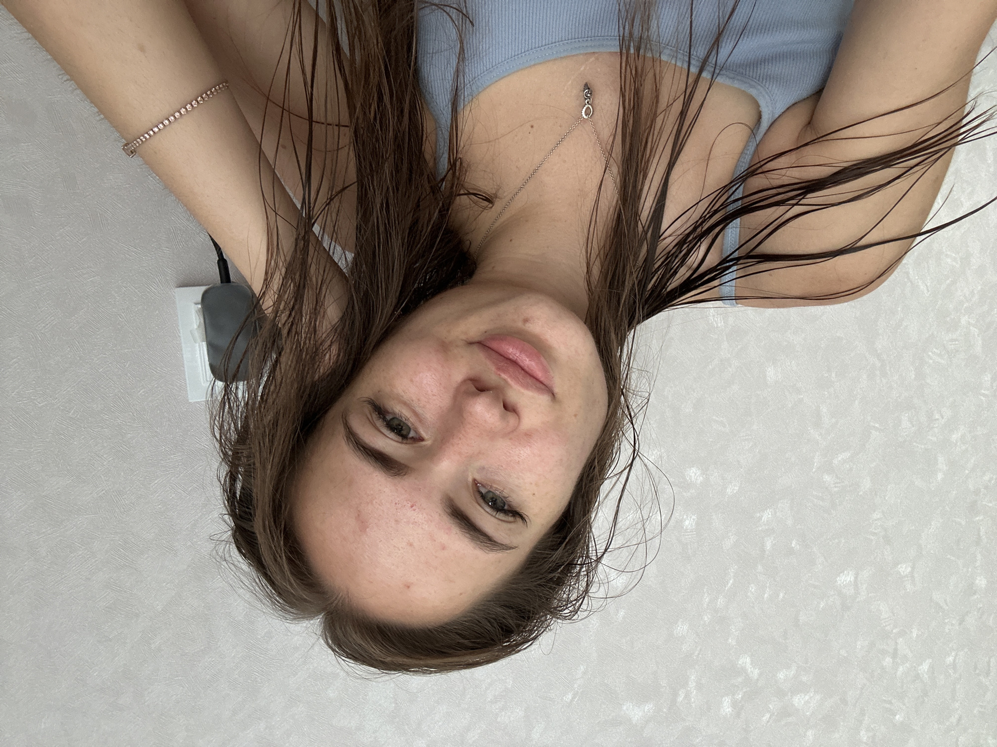 A young woman with wet, brown hair is captured in a close-up, selfie-style photograph. She is wearing a light blue tank top and a delicate silver necklace with a small pendant. Her right arm is resting behind her head, and a sparkling bracelet adorns her wrist. Her skin has a light complexion with some freckles and minor blemishes. Her gaze is directed towards the camera with a gentle smile. The background consists of a textured, light-colored wall. The scene appears to be indoors, in a domestic setting.