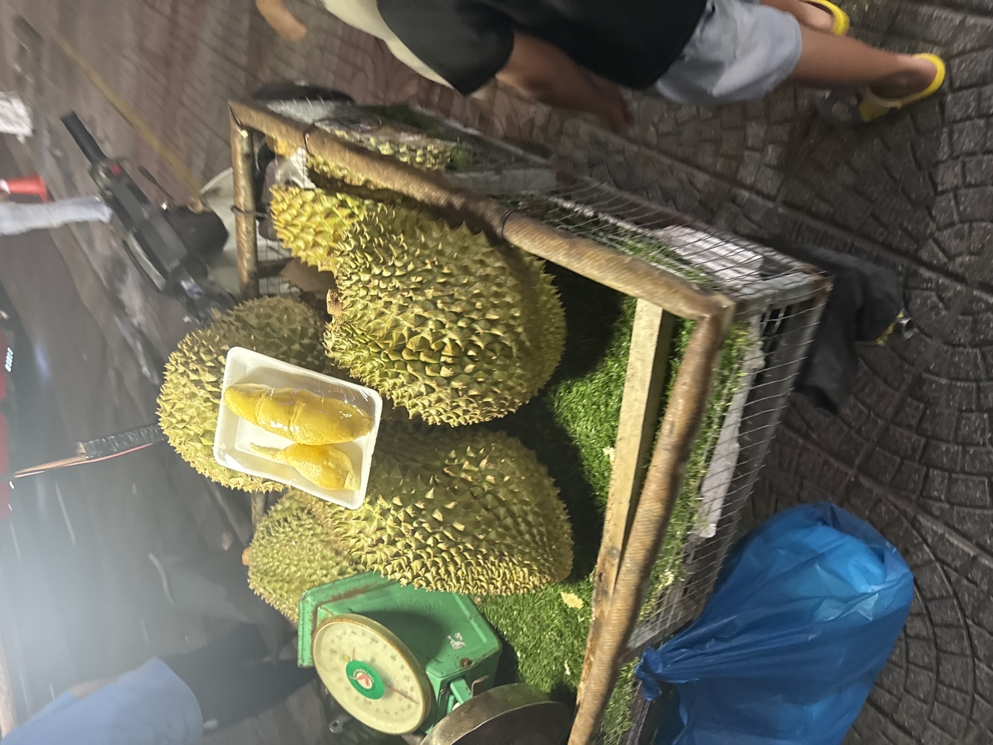 The image depicts a street-side durian stand, likely in Đà Nẵng, Vietnam, based on the prevalence of durian sales in the area. A metal cage contains several whole durians, characterized by their spiky exterior. A scale is positioned beside the durians, used for weighing the fruit for sale. A white styrofoam tray with durian segments is visible, suggesting that pre-cut portions are also available. The foreground shows a blue plastic bag. The upper part of the image shows the legs of people standing around the stand. The ground is bricked.