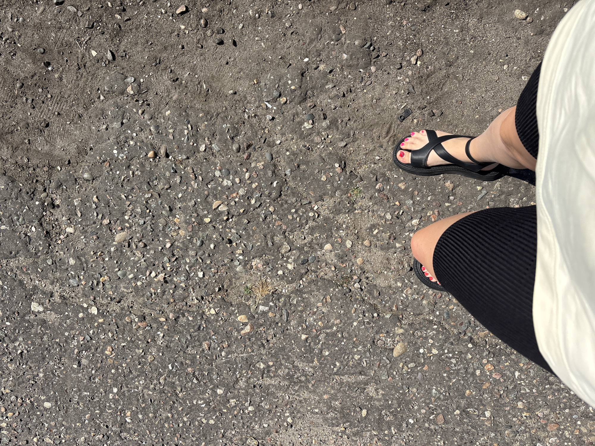 A first-person perspective shot shows a person's legs and feet as they stand on a ground surface composed of dark gravel and dirt. The person is wearing a black skirt with ribbed texture and a white top. Their feet are adorned with bright pink nail polish and black criss-cross strap sandals. The background is an outdoor environment with a ground texture characteristic of a path or unpaved area in a city.