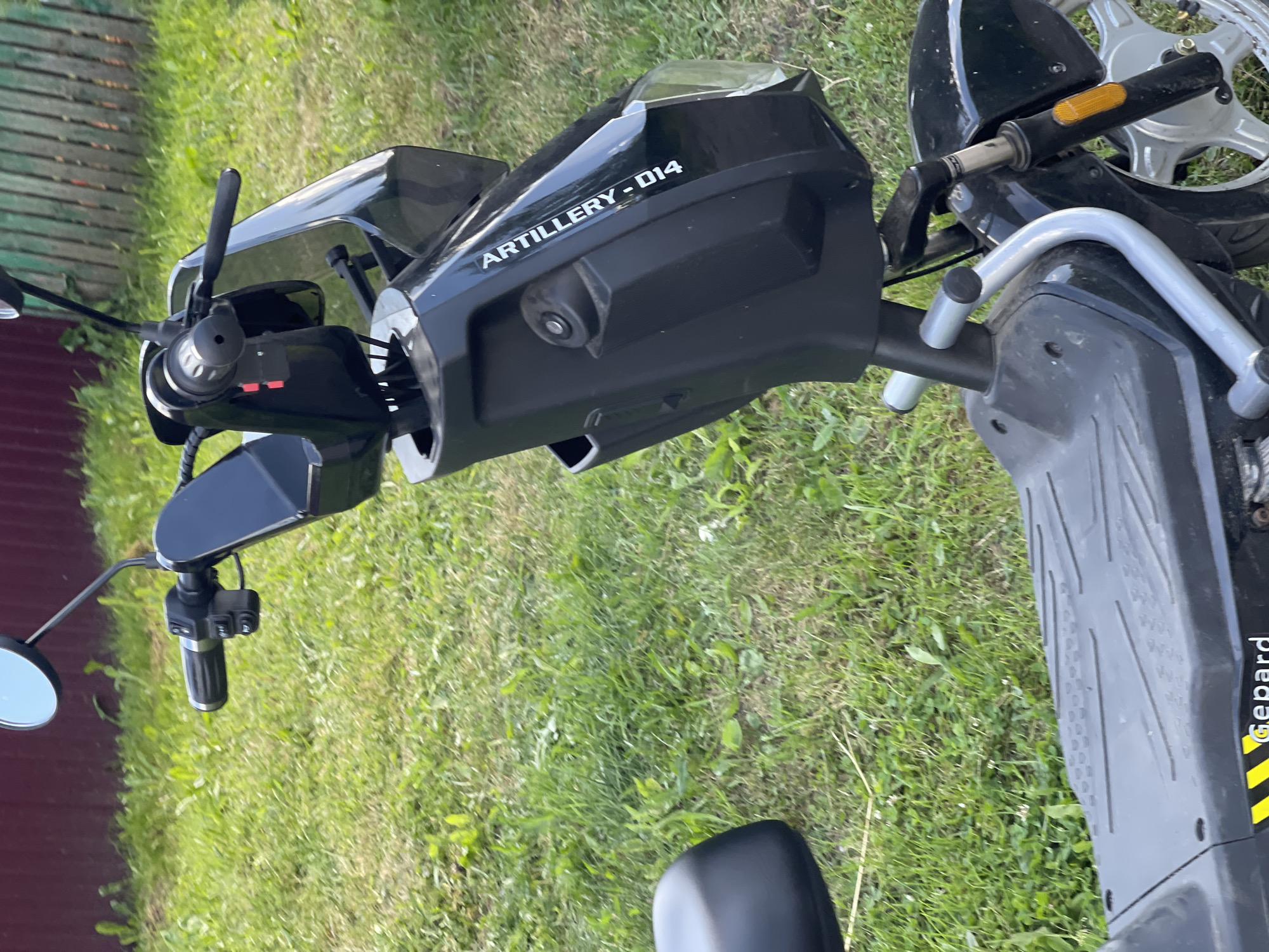 An outdoor medium shot captures a black electric scooter parked on a grassy area. The scooter's handlebars and front portion are visible, with the "ARTILLERY - 014" branding clearly displayed on the side of the front panel. The scooter has a dark seat and a textured footplate. In the background, a red fence and a green wooden fence are partially visible, suggesting a residential or garden setting. The environment appears to be an urban area in Zarechny, Russia.
