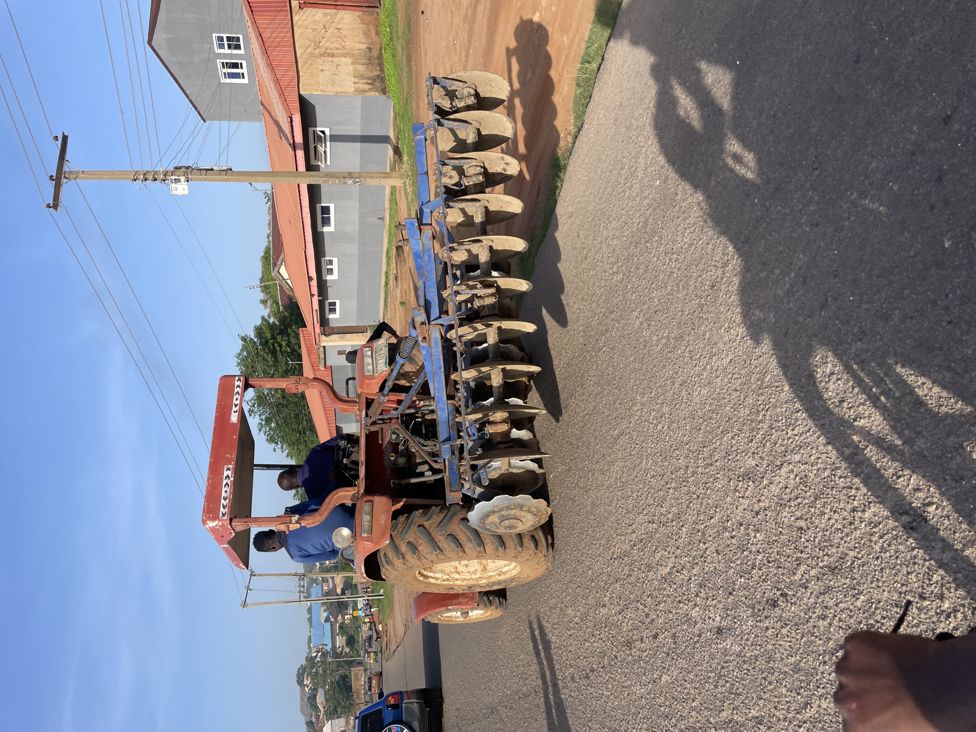 A bright, sunny day in Jos, Nigeria, the scene is dominated by a red tractor with a plow attachment, moving on a paved road. Two men, presumably the driver and a passenger, are seated on the tractor. The tractor's rear tires are caked with mud, suggesting recent use in a muddy environment. The plow, a heavy piece of agricultural equipment with multiple metal discs, is attached to the rear of the tractor. In the background, modern buildings with grey facades and red roofs, along with power lines on wooden poles, suggest an urban or semi-urban setting. The road appears to be a main thoroughfare with other vehicles visible in the distance, indicating ongoing traffic. The overall impression is a functional scene of agricultural work taking place within a developed area.