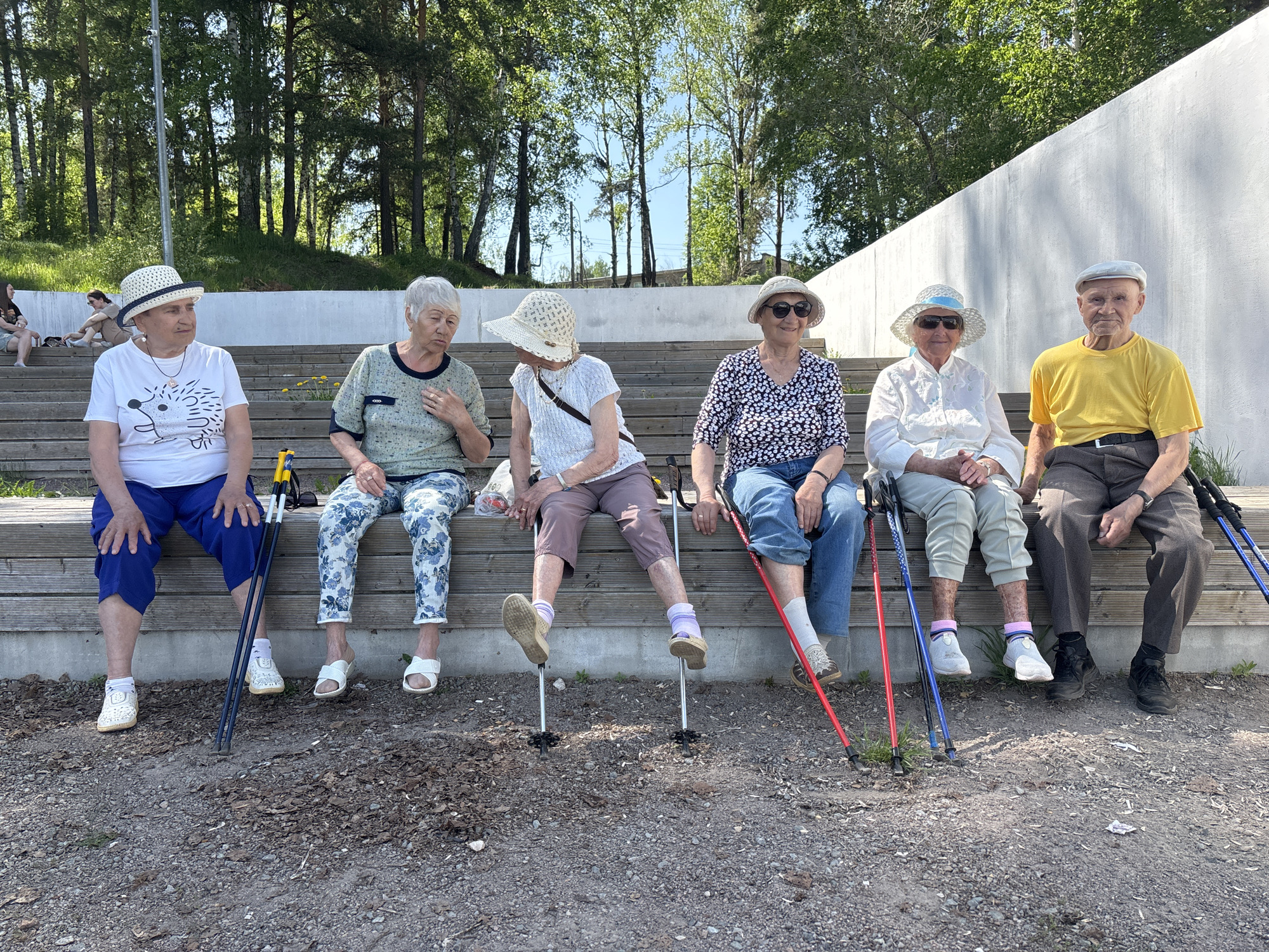 Six elderly individuals are seated on a wooden bench in an outdoor setting. Each person has walking poles. The backdrop includes trees and a white wall. The setting is likely a park or recreational area in Chaikovsky, Russia. The ground is covered with gravel and scattered leaves. The individuals appear to be resting, likely after an exercise or walking activity.