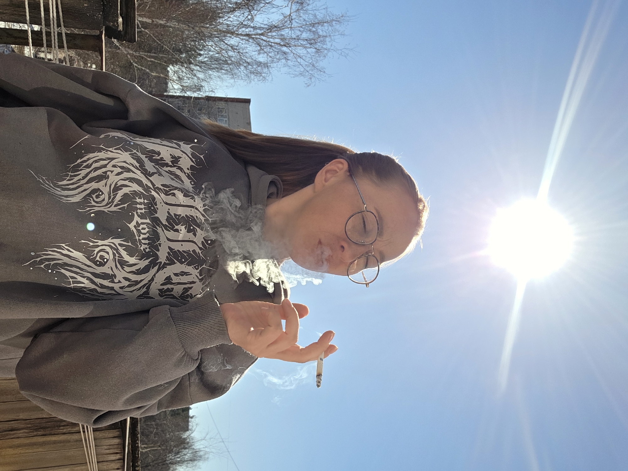 A woman with reddish-brown hair and wearing round glasses is smoking a cigarette outdoors on a sunny day. She is exhaling a cloud of smoke. She is wearing a gray hooded sweatshirt with a stylized white design on the front. The background consists of a bright blue sky with a strong sun, a bare tree, and part of a building. The location is Chaikovsky, Russia. Wooden fencing is partially visible.