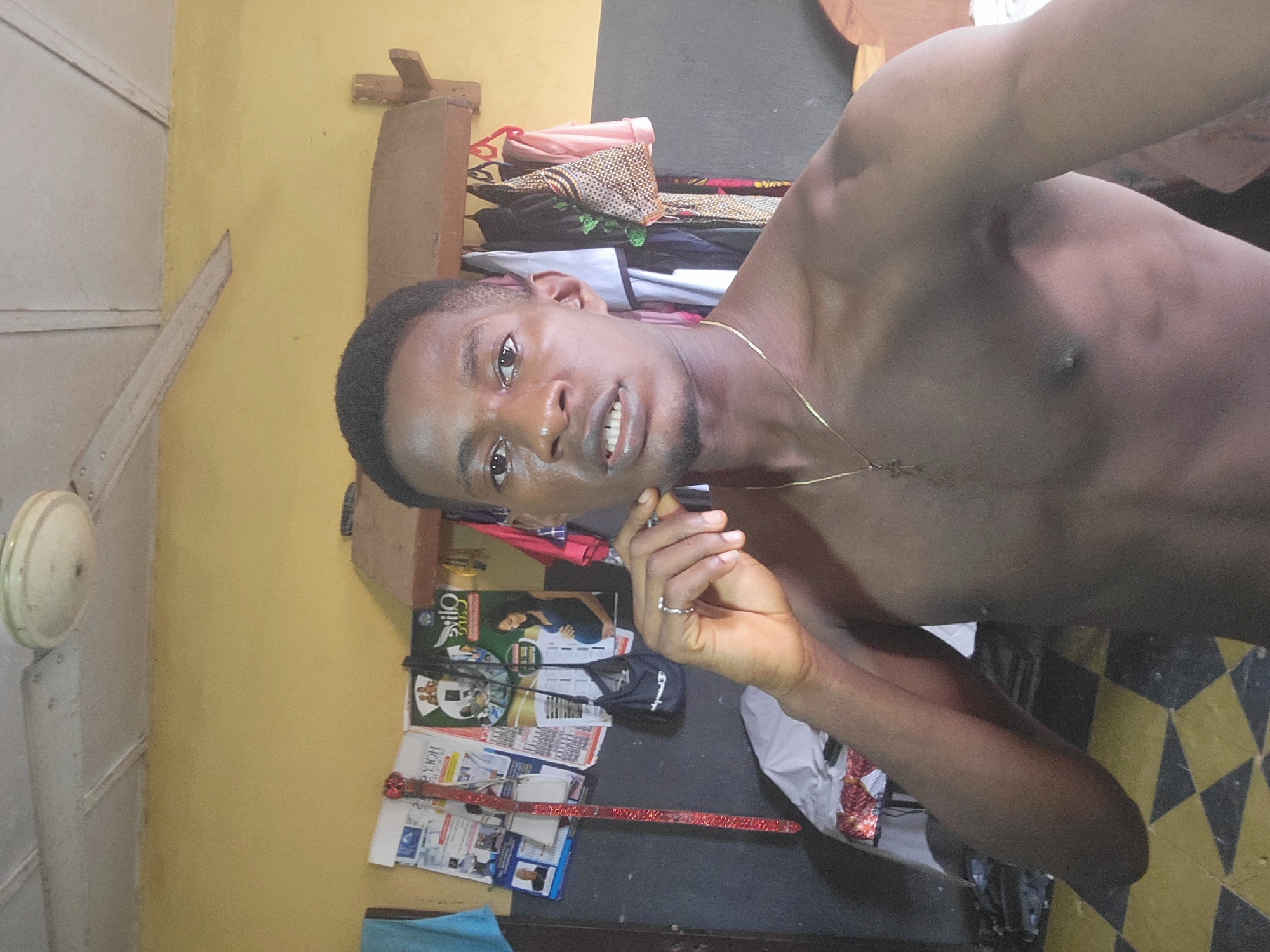 A young man is pictured from the chest up, looking directly at the camera. He is shirtless and wearing a thin gold chain with a cross pendant. His right hand is raised to his face, with his ring finger touching his chin. He has dark hair and appears to be in a room with yellow walls and a tiled floor. There are posters and clothing visible in the background. A ceiling fan is mounted on the ceiling. The image appears to be a selfie.