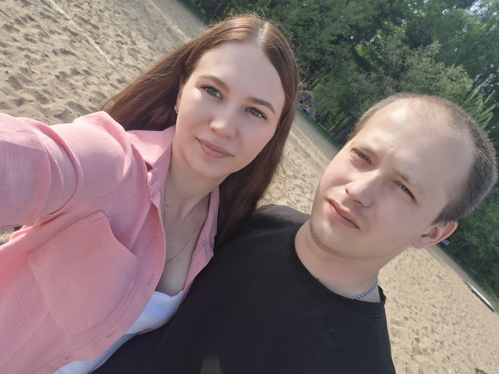A young couple poses for a selfie outdoors. The woman has long brown hair and wears a pink shirt over a white top. The man wears a black t-shirt and a silver chain. They are standing on a sandy surface, possibly a beach. In the background, there are trees, suggesting a park or wooded area. The location is Noviy, Russia.