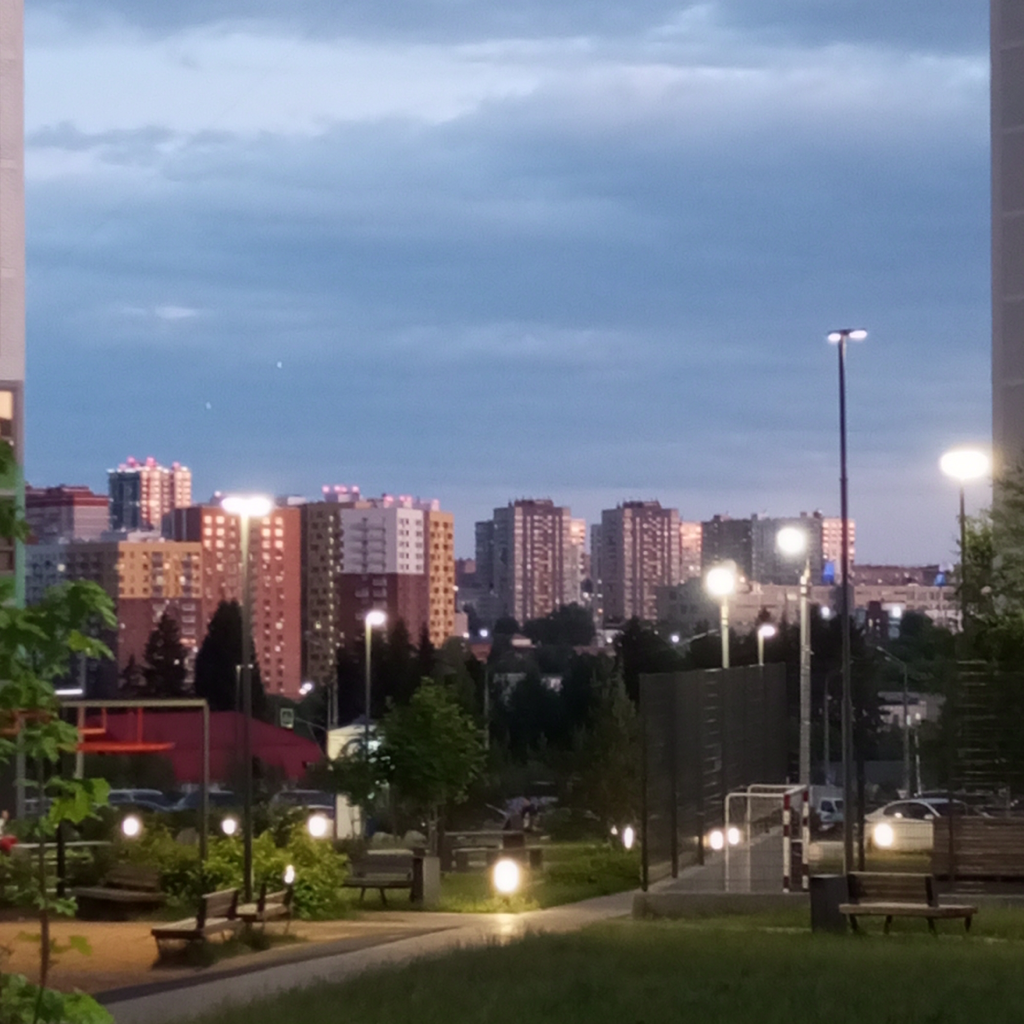 This is a dusk shot of a residential area in Izhevsk, Russia. The image showcases a cityscape dominated by multi-story apartment buildings, some illuminated by interior lights. In the foreground, a park area is visible with benches, green grass, and trees. Streetlights illuminate pathways and open spaces. A basketball hoop structure with a red backboard is also present. The sky is overcast with muted blue and grey tones. The scene depicts a typical urban residential environment in the early evening.