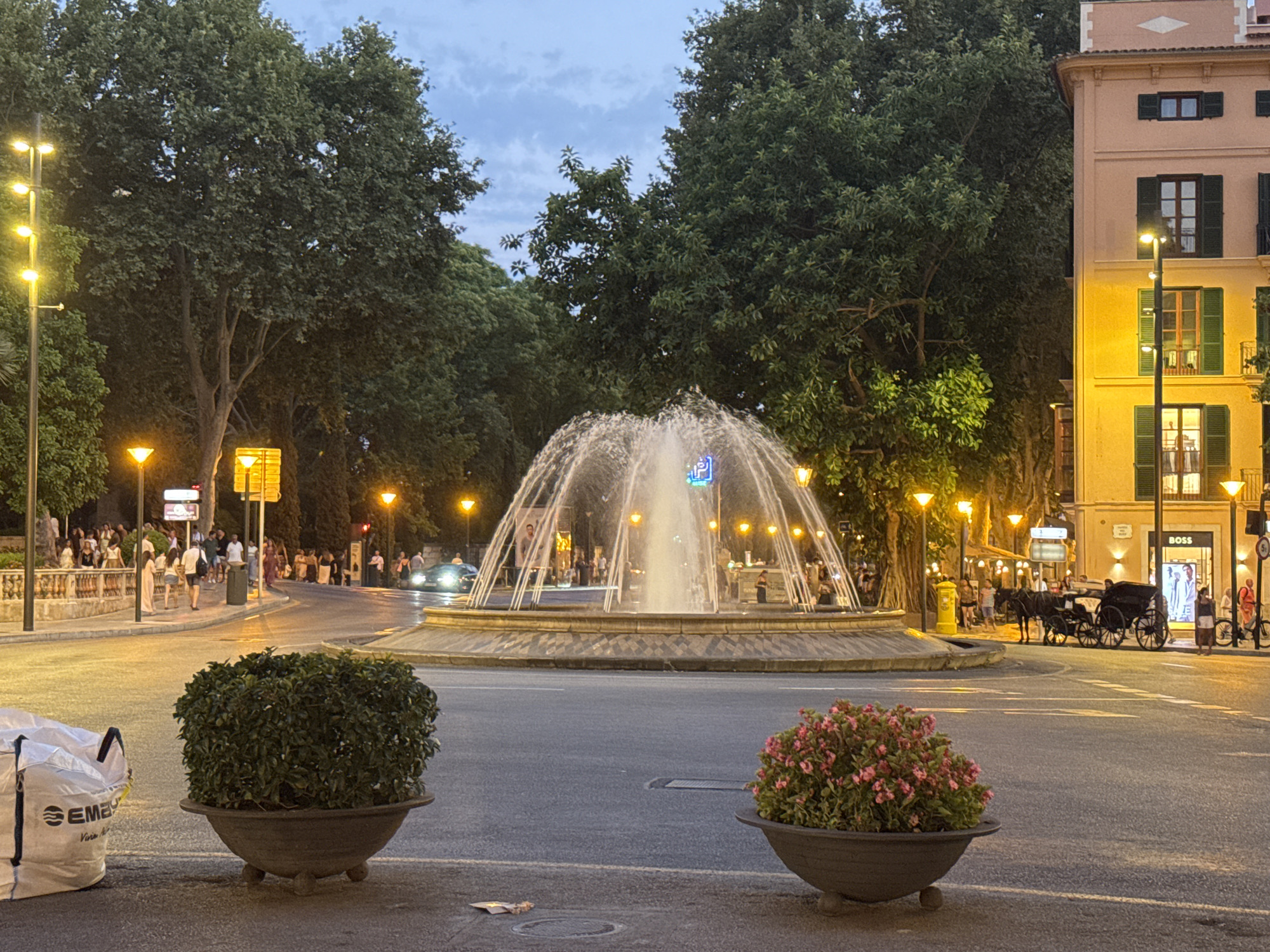 The image depicts a street scene in Palma, Spain, featuring a prominent fountain in the center of a roundabout. The fountain is actively spraying water, illuminated by overhead lights. Surrounding the roundabout are mature trees, providing a lush green canopy. On the right side of the frame, a building with a yellowish facade and green shutters is visible, housing a "BOSS" store. Several street lamps with yellow lights are strategically placed throughout the scene, illuminating the pathway and surrounding area. In the foreground, two large planters are present: one on the left containing a dense green bush, and another on the right with a vibrant display of pink flowers. In the background, pedestrians are seen walking along the street and near the fountain. A horse-drawn carriage is parked on the right side of the roundabout, and a car is visible driving through the intersection. A large white bag with "EMEP" printed on it is positioned in the lower left corner of the image. The sky is a muted blue with scattered clouds, suggesting it is either early morning or late evening.