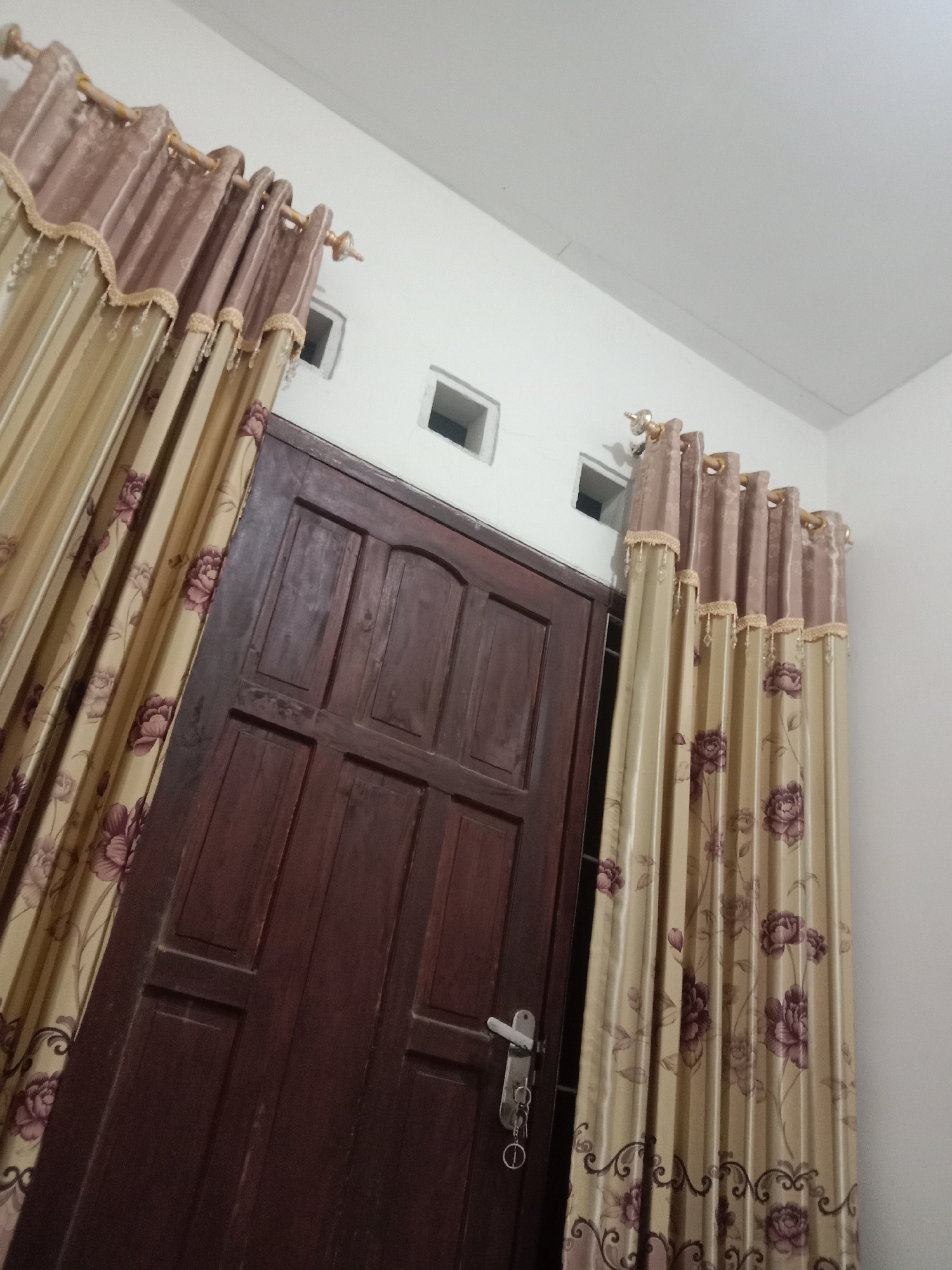 The image shows an interior view of a room, possibly in Brangsong, Indonesia. The main focus is a dark wooden door with a metal doorknob and a set of keys hanging from it. Flanking the door are two sets of decorative curtains. The curtains on the left and right are identical, featuring a floral pattern of large purple flowers and green leaves on a beige background. The top of the curtains has a darker, possibly velvet, trim with gold beadwork and tassels. Above the door, there are two small, square ventilation grates in the white wall. The room appears to be lit by ambient light, casting soft shadows.