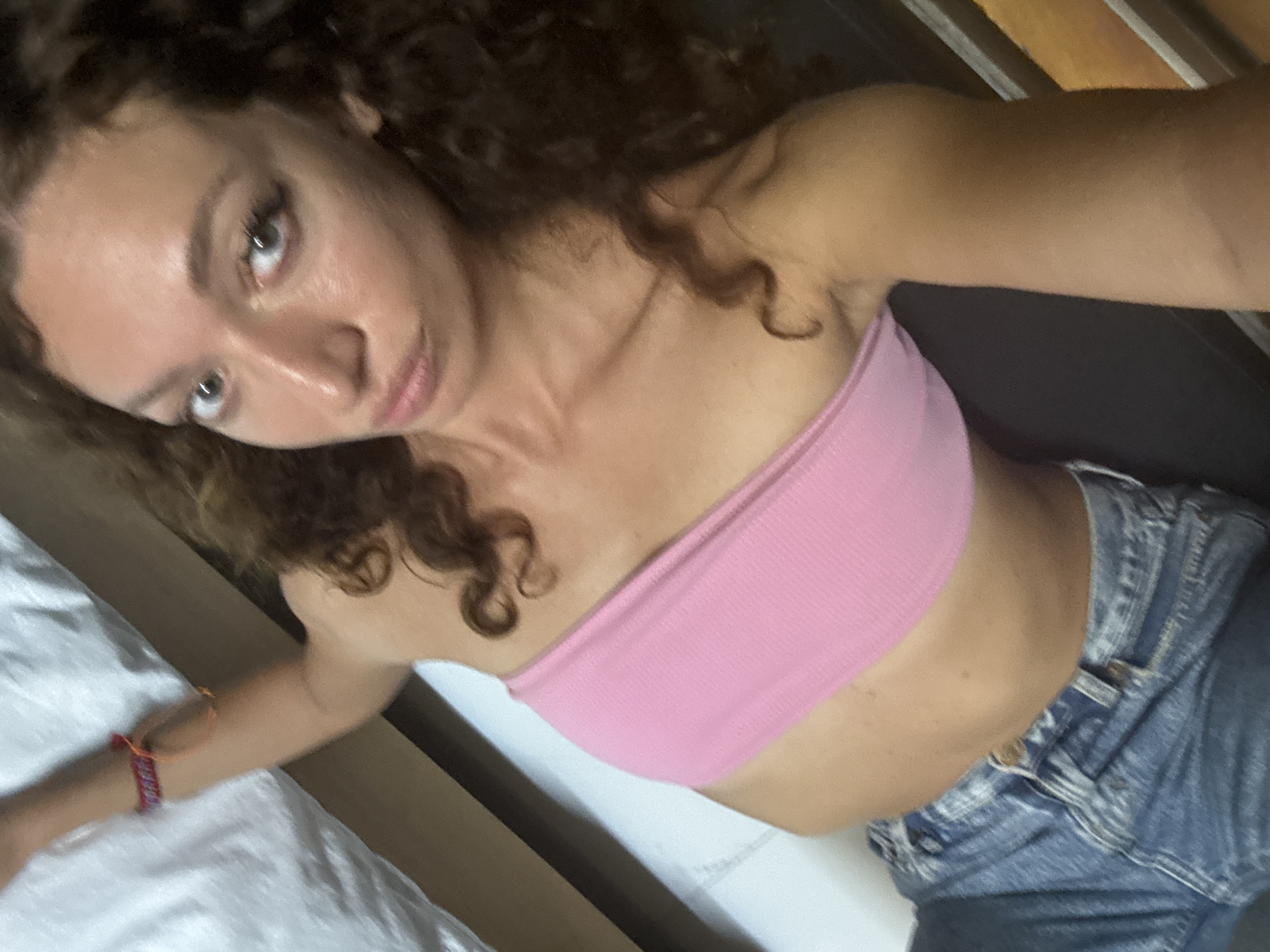 A woman with curly hair and fair skin is posing for a selfie. She is wearing a pink tube top and blue denim jeans. She is lying on what appears to be a white bed. She is wearing a multi-colored bracelet on her left wrist. The location is unknown and there is no information to suggest this image was taken in Than Uyên, Vietnam.
