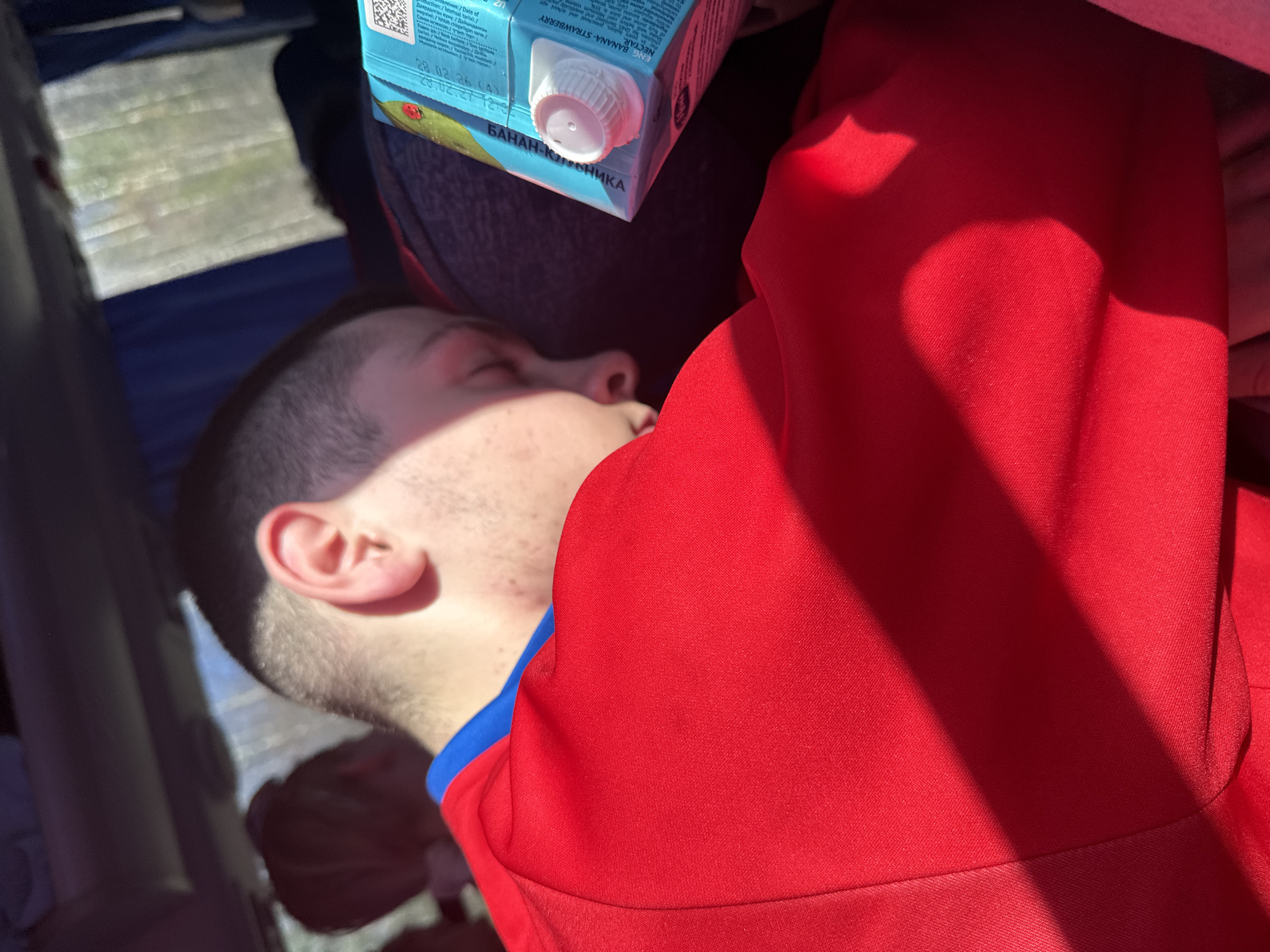 The image shows a person sleeping indoors. The person is lying down, wearing a red shirt with a blue collar. A carton of juice is resting near their head. The text on the carton indicates that it is "Banana-Strawberry" juice. The OCR text implies it might be produced in Uzbekistan (UZ) and intended for the preschool market. The image contains text with dates 28.02.26 and 28.02.21 12:3. The image's location, Челва, Russia, is not visible.
