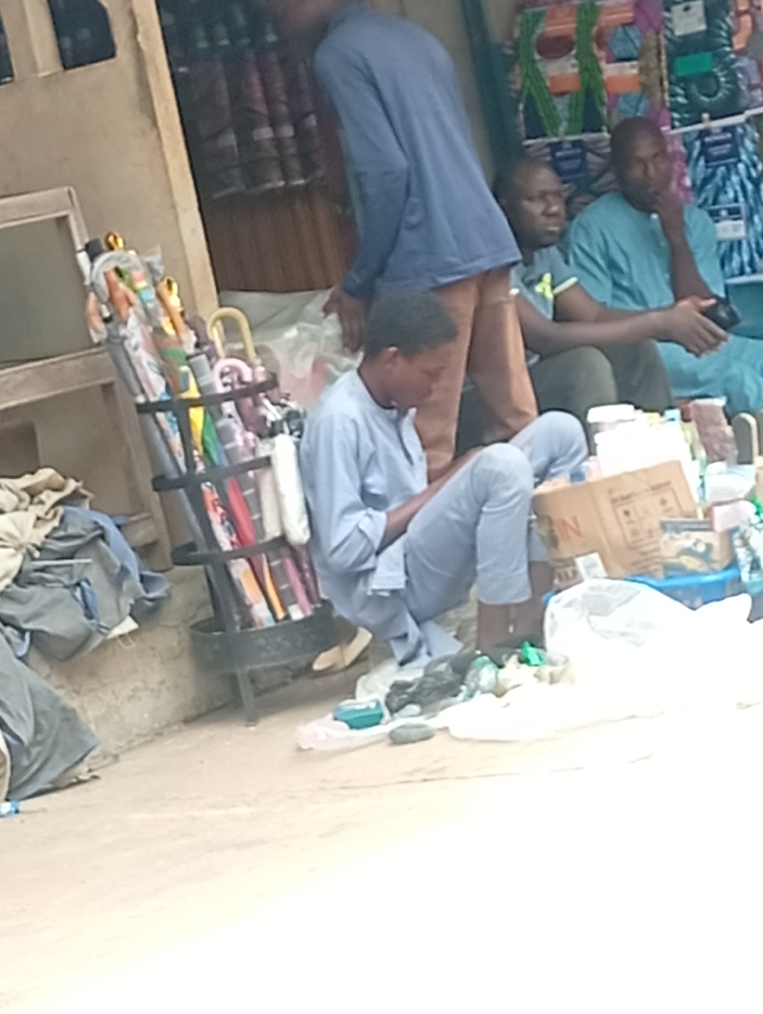The image depicts a street scene in Maiburiji, Nigeria. A young man in light blue clothing sits on the ground amidst an array of goods, possibly engaging with a mobile device or sorting through items. A display of umbrellas stands nearby. Two men are seated in the background, one wearing a light blue shirt and the other a green shirt. The ground is littered with various objects, and merchandise is piled around the seated men. A building is visible on the left.